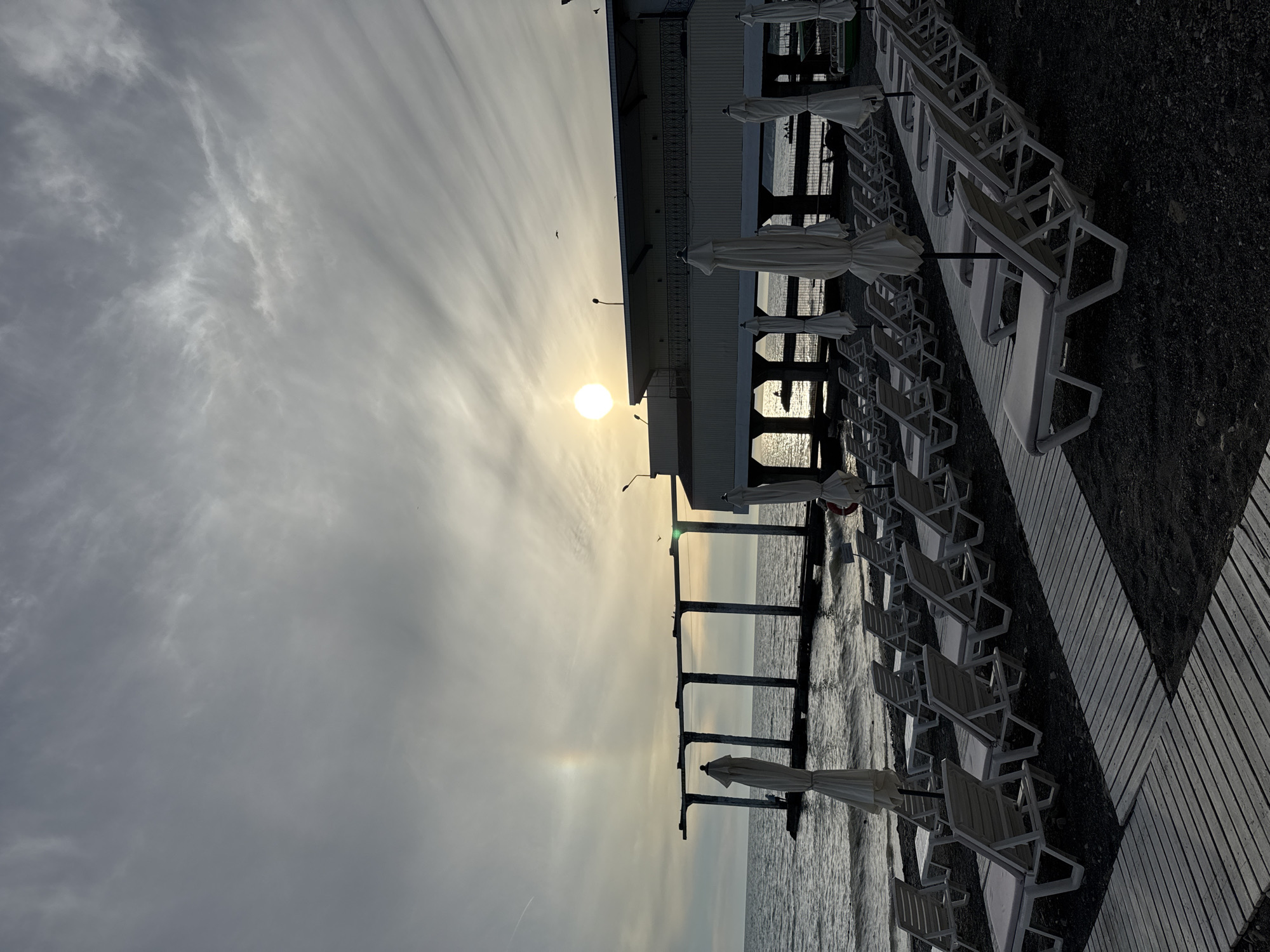 The image depicts an empty beach with rows of white lounge chairs and closed umbrellas. Wooden pathways are laid out on the dark sand. In the background, a pier extends into the choppy sea. A building with a balcony sits behind the pier. The sun is low in the sky, casting a soft glow, and a halo is visible around it. The sky is overcast with wispy clouds. The overall scene suggests a quiet, possibly early morning or late evening on a beach in Sochi, Russia, given the architectural style and coastal setting. There are no people visible in the immediate foreground, but a few figures can be seen on the pier in the distance.