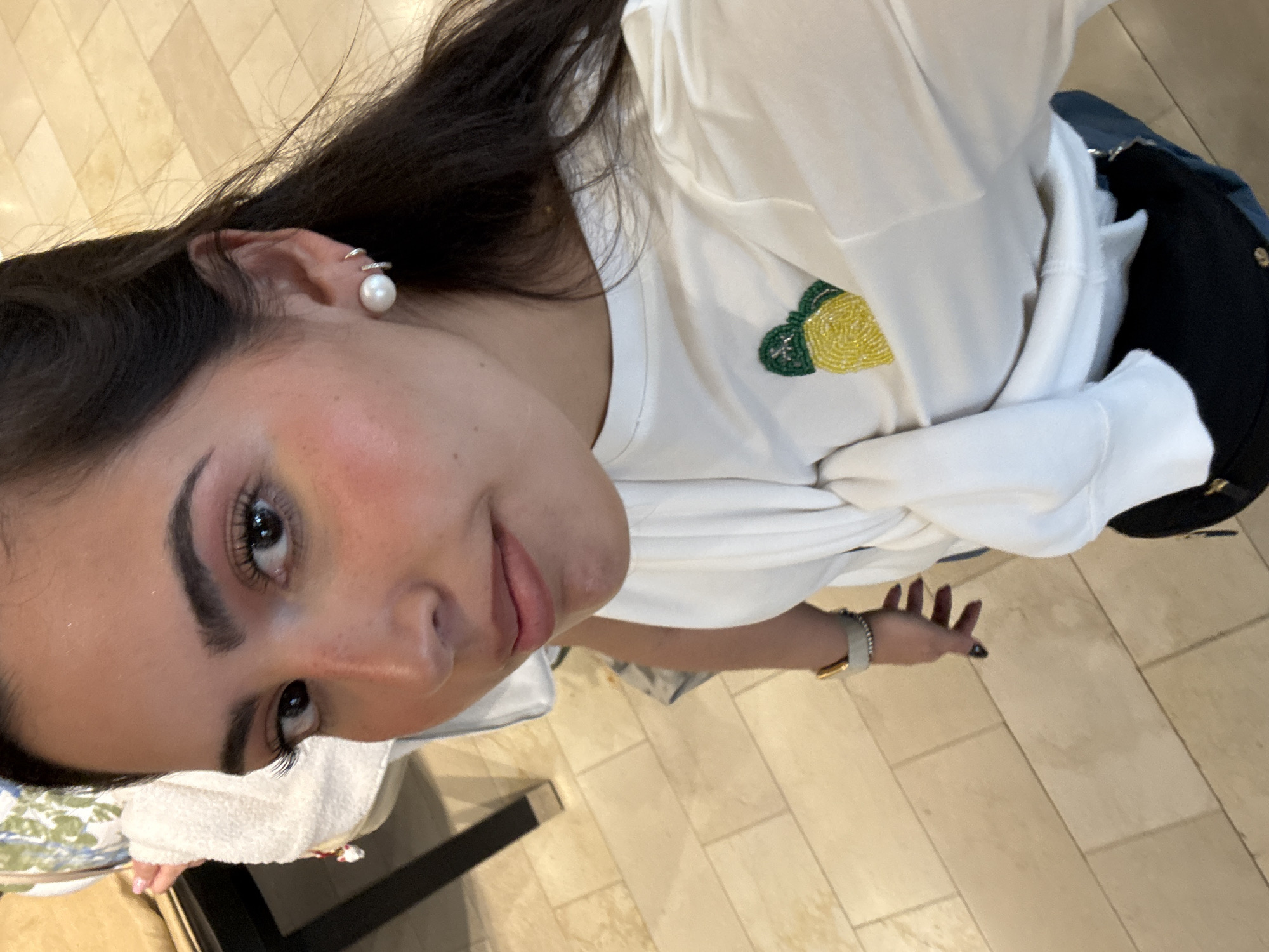 A woman is depicted from a high-angle perspective indoors with tiled flooring. She has dark hair, fair skin, and is wearing a white shirt adorned with a beaded pineapple design. A pearl earring is visible on her left ear. Her left hand is partially visible wearing a light-colored watch, with her fingernails painted dark. She is seated on a chair, with a white towel on her lap. The background includes a black object that appears to be part of her belongings. The location is determined from the flooring and general ambiance to be in San Antonio, United States of America.