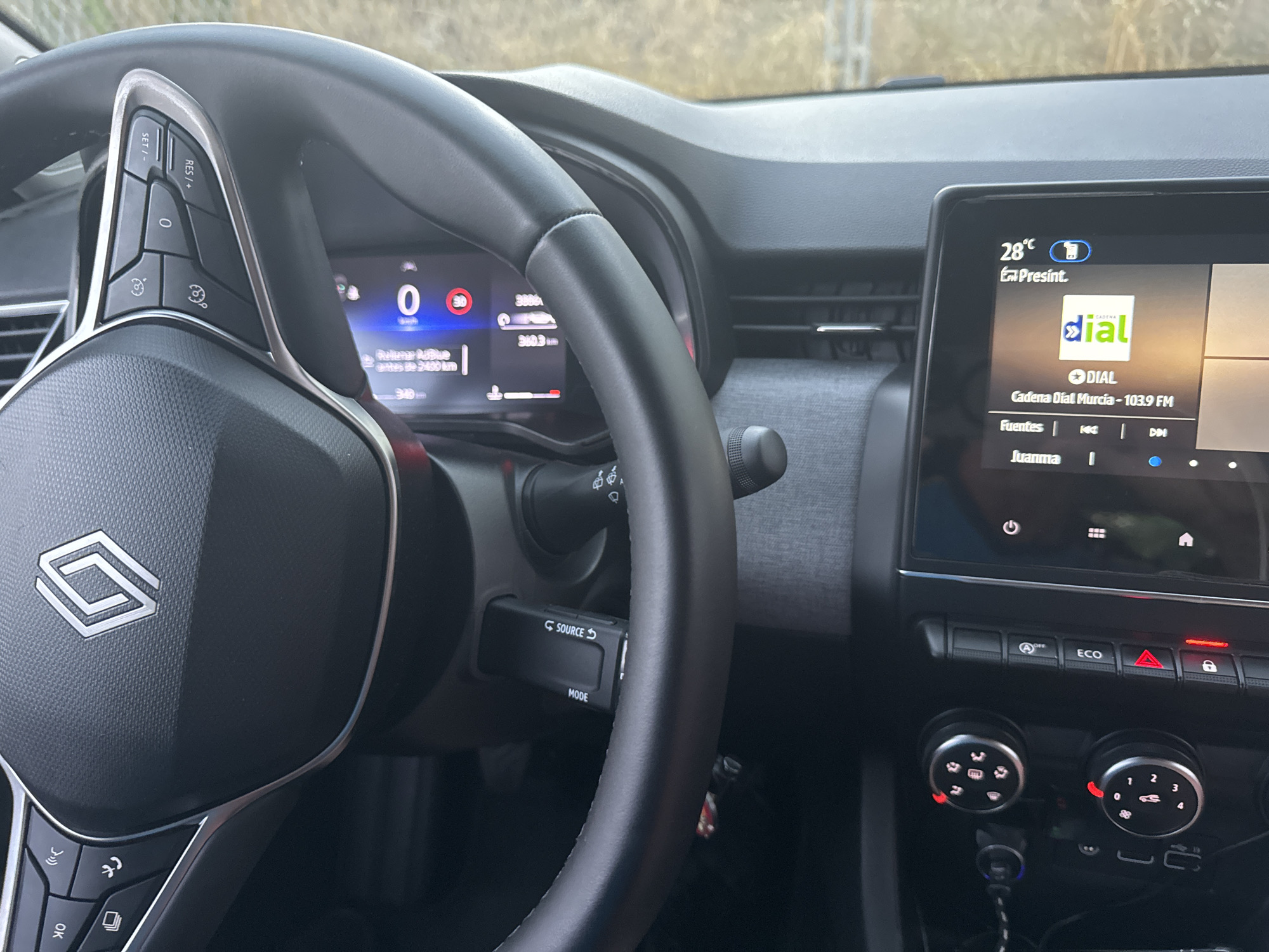 The image shows the interior of a car, focusing on the steering wheel and the central infotainment screen. The steering wheel is black with a silver trim and features several buttons, including controls for cruise control and audio. The infotainment screen displays the current temperature as 28°C and the radio station "Cadena Dial Murcia - 103.9 FM". The car is likely in motion, as indicated by the display of speed and distance traveled on a smaller screen behind the steering wheel. The background suggests an outdoor environment, possibly a road, as seen through the windshield. There are no people visible in the image. The overall scene depicts a common driving scenario in a modern vehicle.