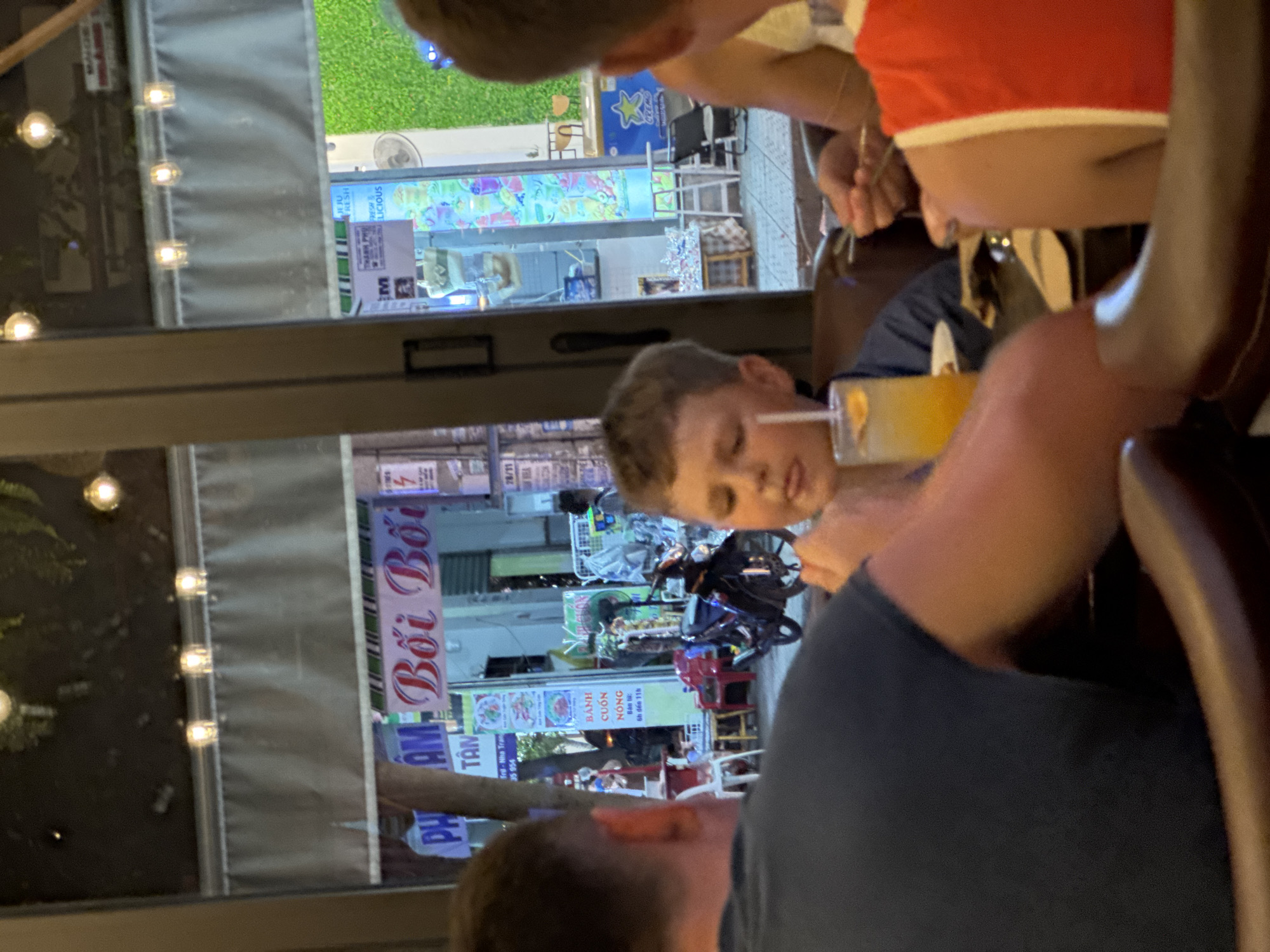 This is a medium-shot, eye-level, indoor photograph taken at night in what appears to be a street-facing restaurant in Nha Trang, Vietnam. The scene is dimly lit with artificial lighting. In the foreground, a young boy with light brown hair is seated at a table, looking towards the camera and sticking his tongue out slightly. He has a glass of orange beverage with a straw and an orange slice in front of him. To the left, the back of an adult male wearing a dark blue shirt is visible, as is the arm and shoulder of another person in an orange and white top to the right. The background shows a street scene through a large window, with storefronts displaying various signs, including one that reads "Bồi Bồi" in red lettering. There are also lights, a motorcycle parked outside, and green decorative foliage. The overall ambiance suggests a casual dining setting on a busy street.