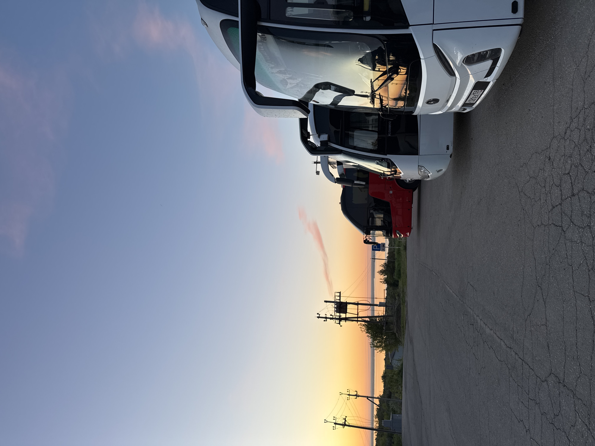 The image shows a row of buses parked in a parking lot in Khvalynsk, Russia at sunset. The foreground features the front of several white buses parked close together, with a red bus visible behind them. The background shows the skyline with the sunset casting orange and pink hues in the sky. Utility poles and wires are also visible in the background landscape. The parking lot surface is cracked asphalt.