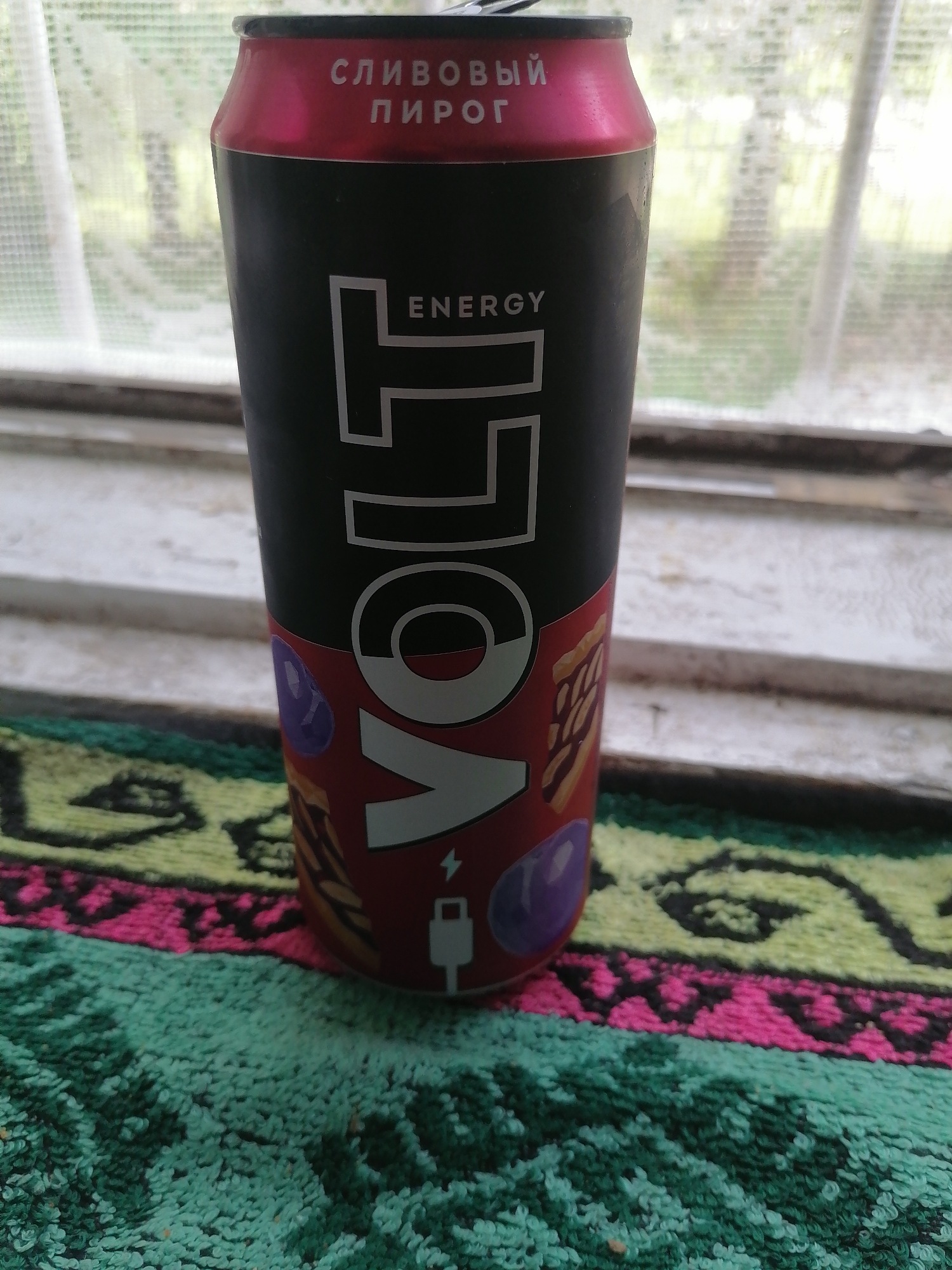 A full can of VOLT energy drink, red and black with white lettering, sits on a patterned rug placed on a windowsill. The rug has a yellow, pink, and green geometric pattern. Behind the can, a window is visible, partially obscured by sheer white curtains. The background outside the window appears to be foliage and trees. The can itself has text that appears to read "СЛИВОВЫЙ ПИРОГ" (Plum Pie) at the top. The VOLT logo is prominent on the can, along with illustrations of what appear to be purple berries and a slice of pie. A charging icon is also visible on the can. There are no people or animals present in the image, and no actions are taking place. The scene is indoors, with the windowsill forming the immediate foreground. The specific location of Большие Ремезёнки, Russia is not determinable from the visual information provided in the image.
