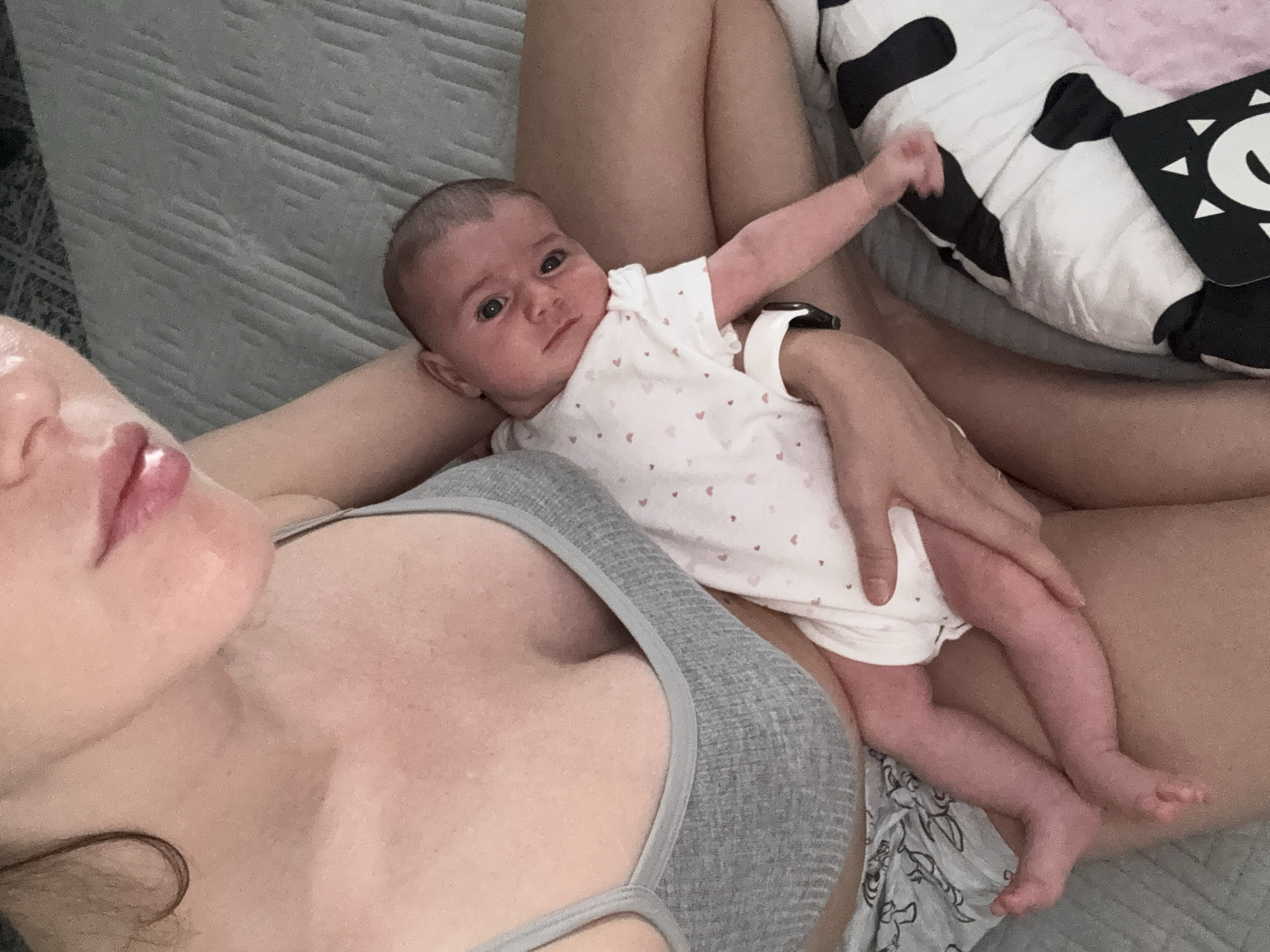 This intimate indoor photograph, set in Isola delle Femmine, Italy, captures a woman relaxing with an infant. The woman is partially visible, with her head, shoulders (wearing a grey tank top/sports bra), and legs in the frame. She has fair skin and pink lips, looking towards the camera. The infant, dressed in a white onesie adorned with small pink hearts, lies comfortably on the woman's lap and stomach. The baby has fair skin and dark eyes, gazing directly at the viewer with a calm, curious expression.
A smartwatch is visible on the baby's left arm, likely worn by the woman. In the background, a light grey textured blanket or bedding is seen, suggesting a bed or couch setting. To the right, next to the baby, there is a black and white cow-patterned pillow or soft toy, and a black and white high-contrast baby card or book with abstract shapes including a large 'C' and triangles. Another patterned fabric is visible beneath the baby's legs. The lighting indicates it is daytime. The scene conveys a moment of peaceful bonding between the woman and the infant. No explicit text is readable within the image.