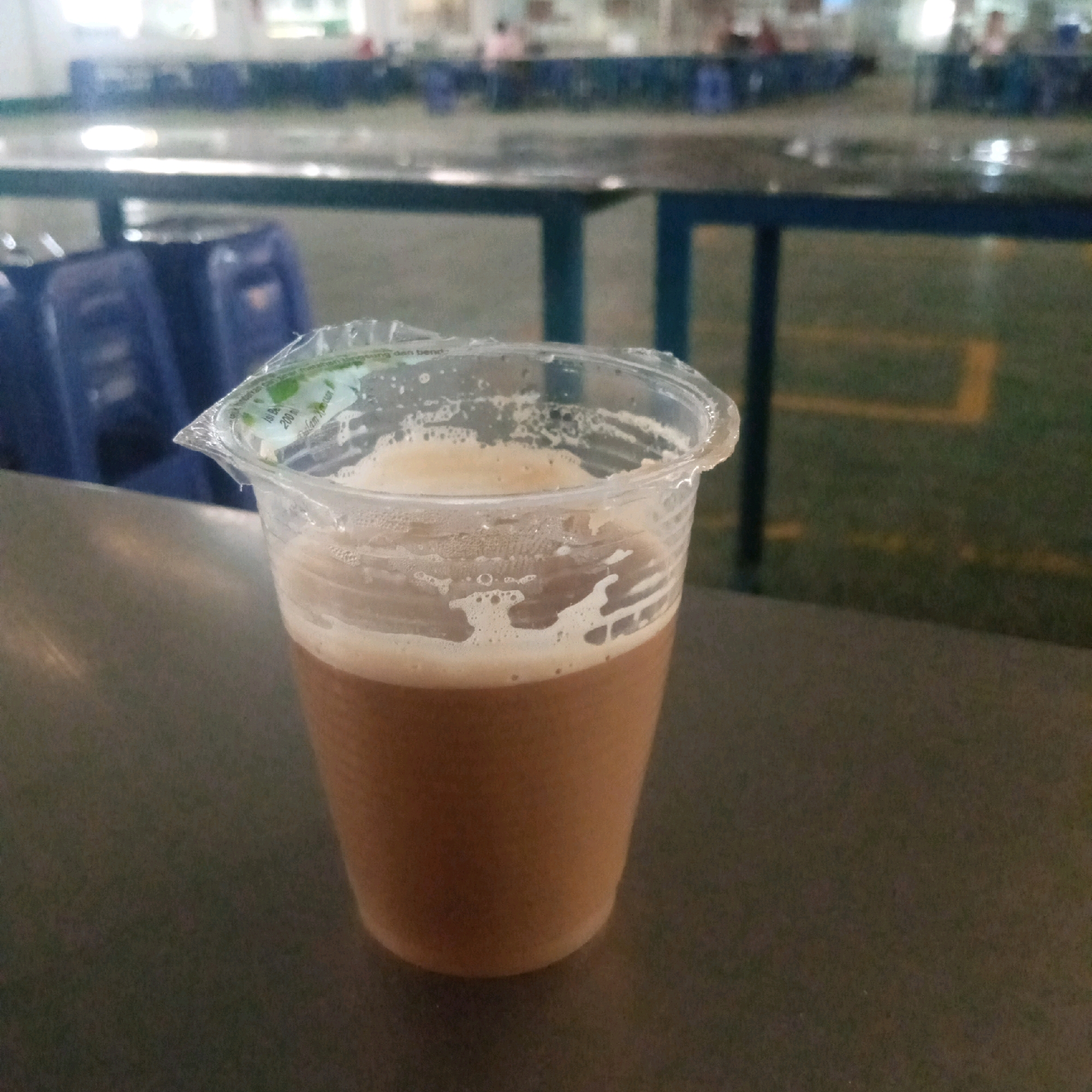 A close-up shot captures a plastic cup filled with a frothy, brown beverage, likely coffee or tea, with foam on top. The cup has a clear plastic lid partially covering it, with some text visible on the lid's edge, including what appears to be "200 ml". The cup is placed on a dark, smooth surface, possibly a table. In the blurred background, rows of blue plastic chairs are visible, some appearing wet. Beyond the chairs, a darker, out-of-focus area suggests an indoor or covered outdoor space, with hints of yellow lines on a green surface. The overall lighting is dim, with some reflections on the surfaces, suggesting it might be evening or an indoor setting with artificial light. There are no people clearly visible or identifiable in the frame. This scene could be at an event venue, cafeteria, or common area in Cijeruk, Indonesia, given the context.