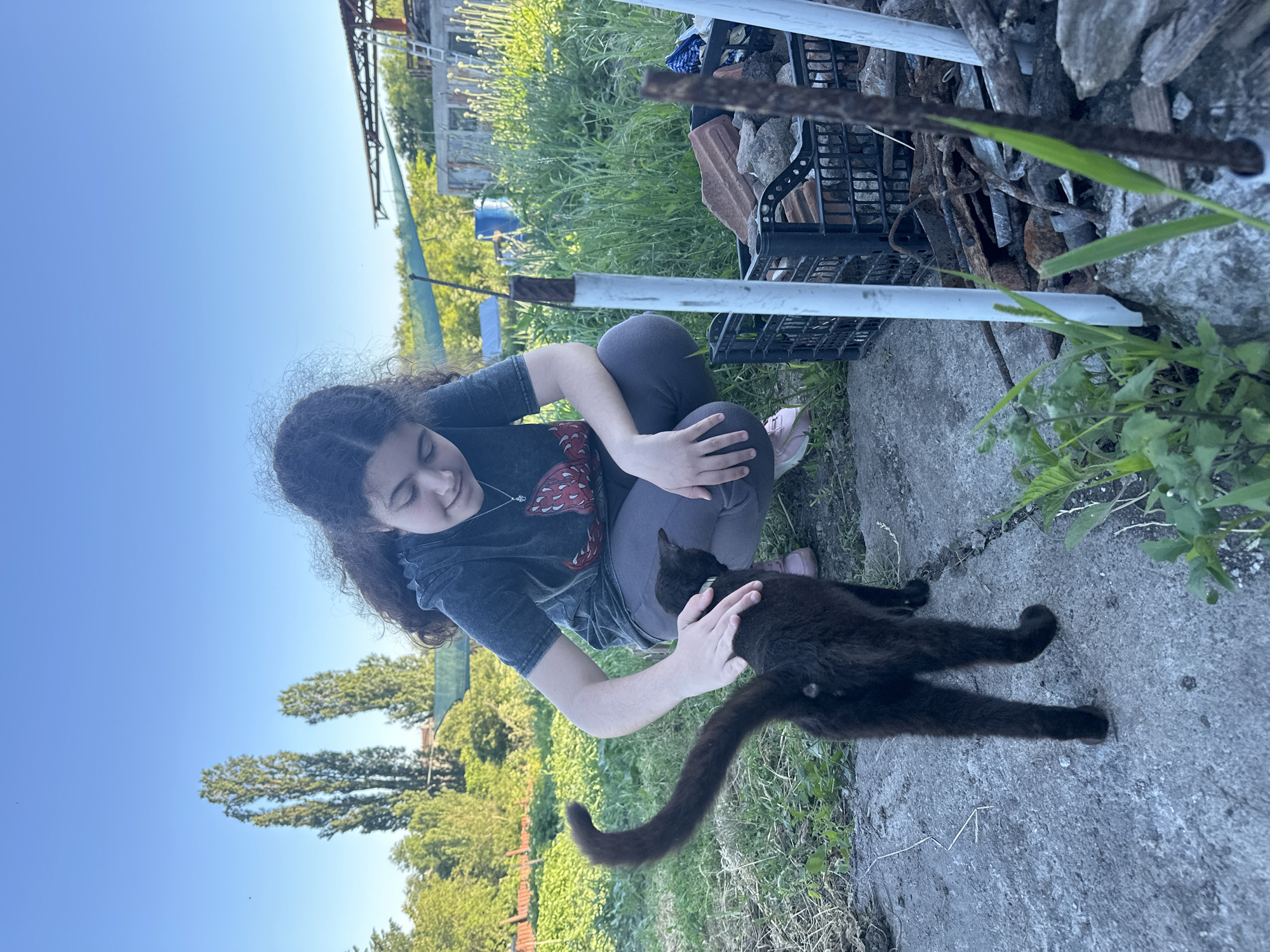 In Ravna Gora, Bulgaria, on a bright, sunny day, a young woman with long, dark, curly hair crouches on a concrete path. She is wearing a dark t-shirt with a red graphic and dark pants. Her right hand gently pets a black cat. The cat is facing away from the viewer, with its tail raised high. The environment is rural, with tall trees and lush green vegetation visible in the background. In the foreground, to the right of the cat, a black plastic crate is filled with bricks and debris, and some rusty metal rods are propped up nearby. The ground is a mix of concrete and dry grass. The sky is a clear, bright blue.