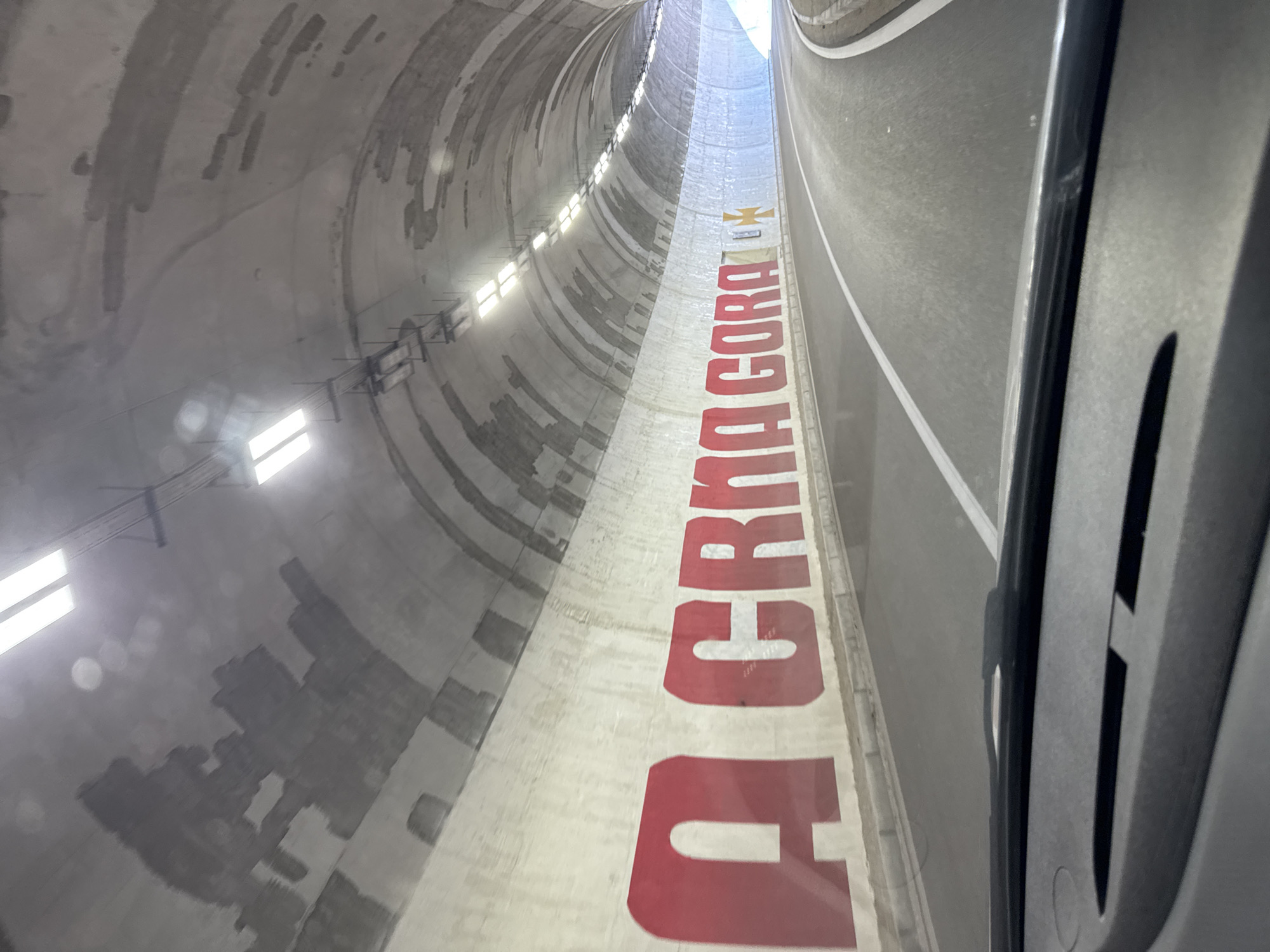 The image shows the interior of a tunnel, likely the Sozina Tunnel, located in Bukovica, Montenegro. Large red lettering on the left wall spells out "CRNA GORA", the Montenegrin name for Montenegro. To the right of the text, there is a yellow cross with four equal arms. The tunnel has a concrete ceiling with visible repairs and lighting fixtures. The road within the tunnel is asphalt with white lane markings. The perspective suggests the photo was taken from the driver's seat of a vehicle. The overall scene is industrial and utilitarian, characteristic of a road tunnel.