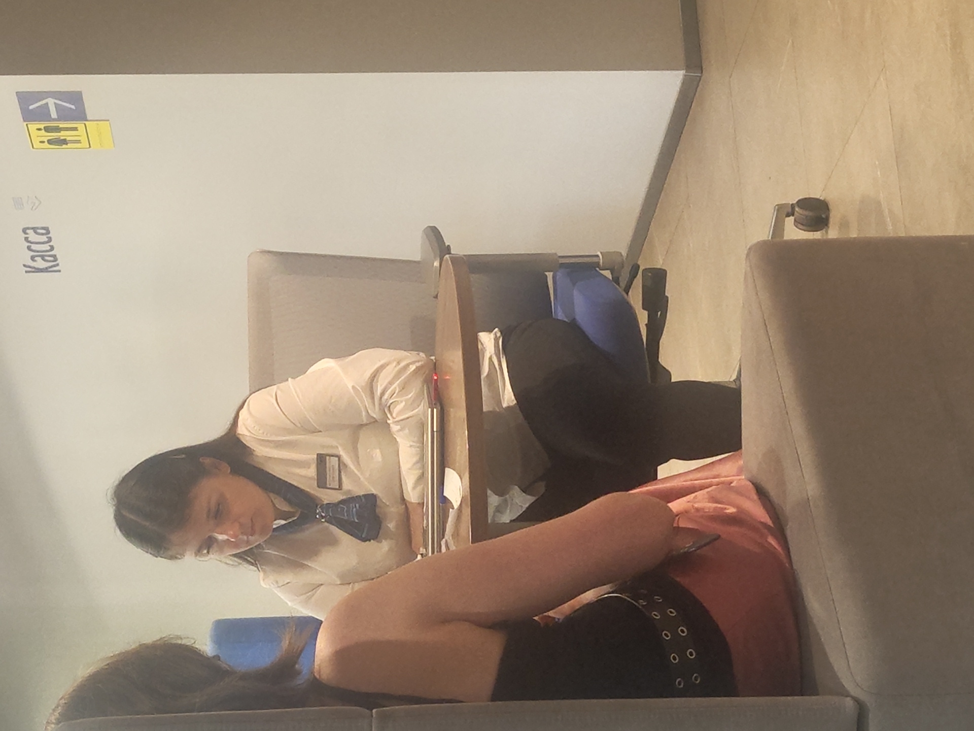 In Ramenskoye, Russia, a woman in a white collared shirt and dark pants sits behind a circular wooden table, facing a seated individual whose back is to the camera. The woman has dark hair, pulled back, and is wearing a blue scarf tied with a knot. A name tag is visible on her shirt. On the table, a laptop and a white piece of paper are present. To the right of the woman, a sign on the wall reads "Касса" (Kassa), indicating a ticket office or checkout. A yellow and blue directional sign with figures of a man and woman, and an arrow pointing right, is also on the wall. The environment appears to be an indoor waiting area or service counter, with a grey upholstered chair to the right of the woman. The floor is tiled in a light brown color.