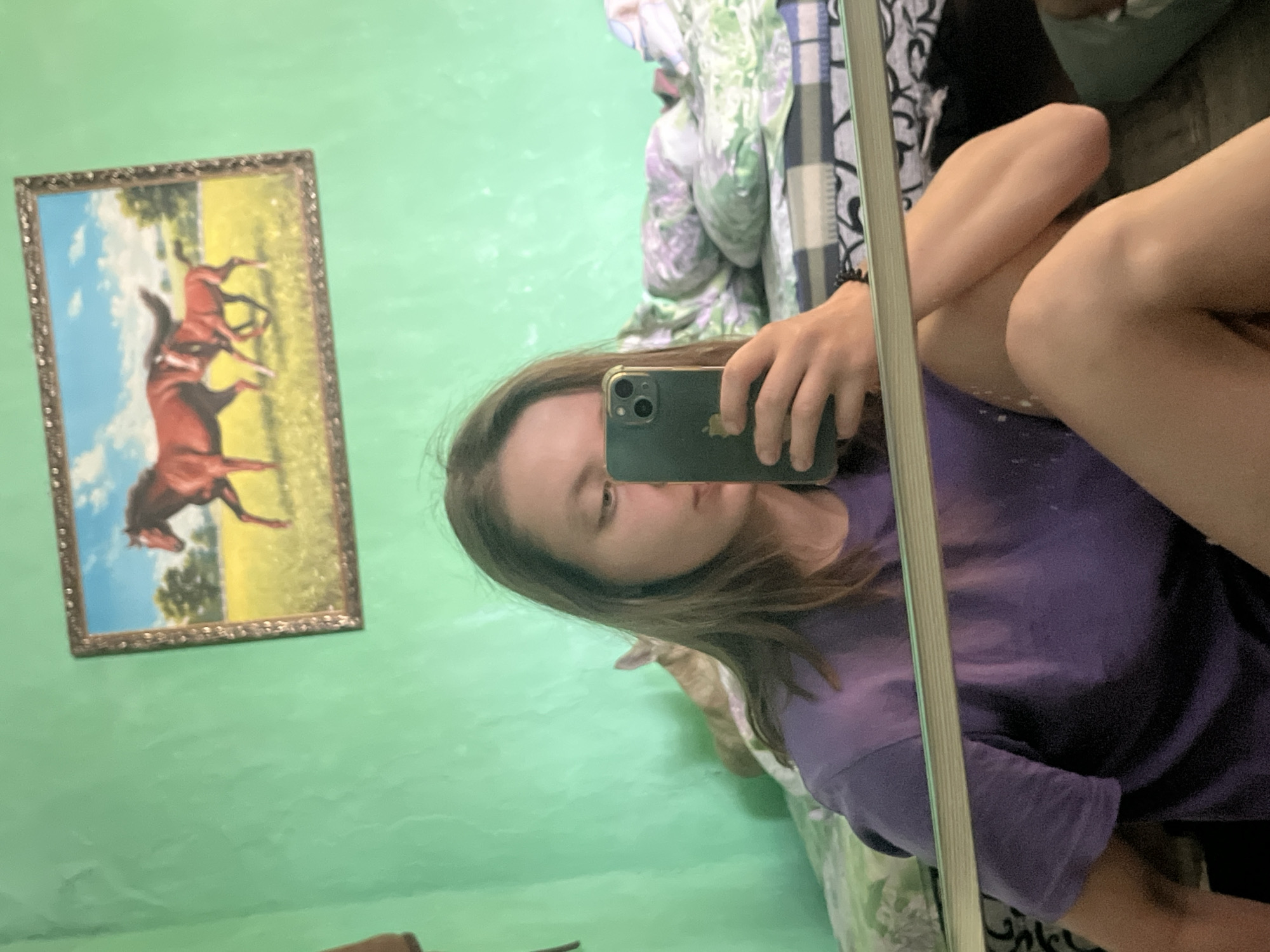 The image is a selfie taken by a young woman in a room. She is wearing a purple t-shirt and has long brown hair. She is holding a smartphone in her right hand, taking the photo. Behind her, a bed with patterned bedding is visible, and a painting of horses hangs on a green wall. A cat can be seen behind the woman, with its ears and head poking up from the bed. The room appears to be a personal space, possibly a bedroom. The location is not identifiable as Nizhny Tagil, Russia.