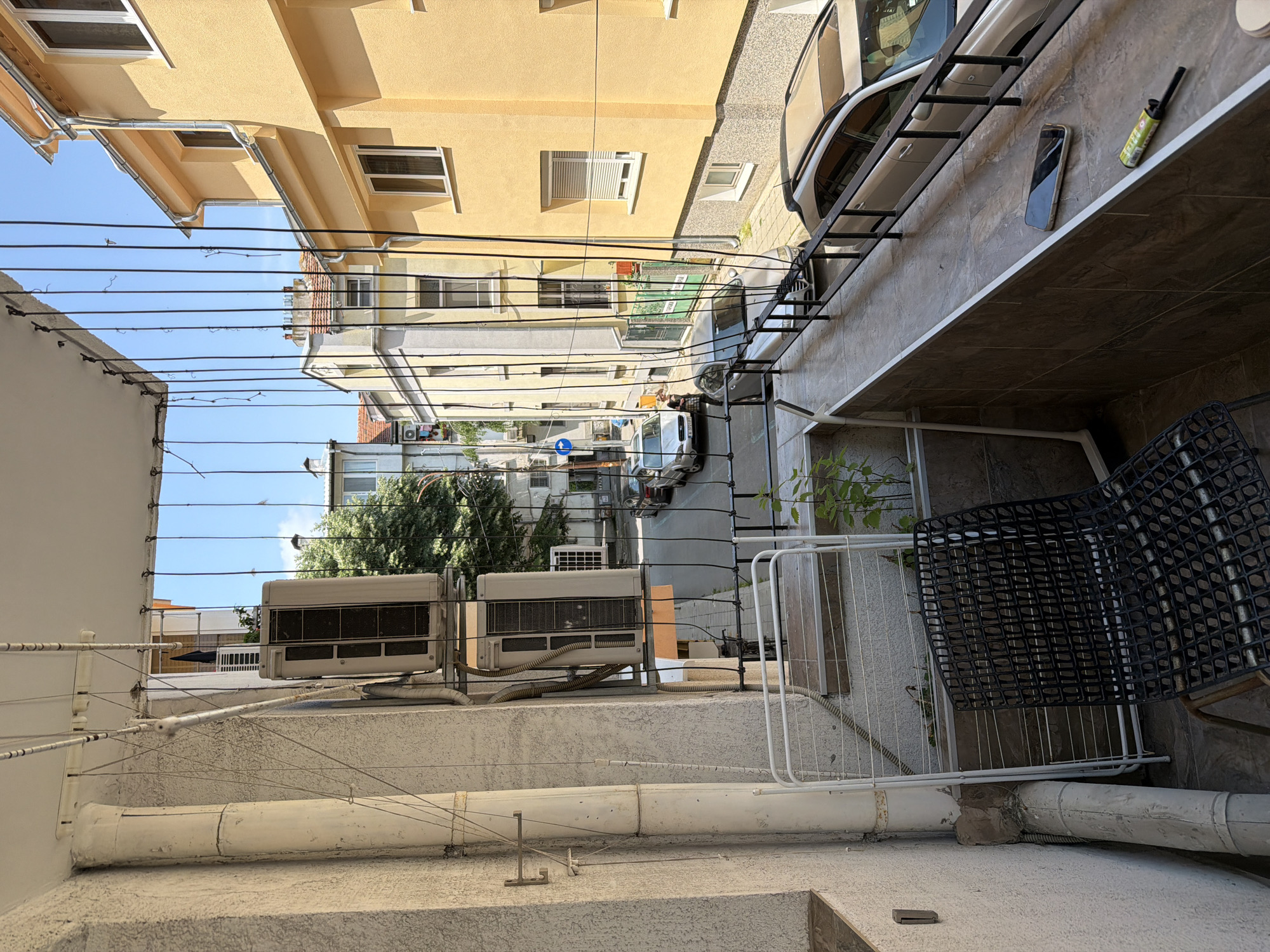 The image, taken from an elevated, rotated perspective, depicts a sunlit urban alleyway or narrow street, likely in Burgas, Bulgaria. The view looks down into the street from a building or balcony.
In the immediate foreground on the right, a tiled terrace or balcony features a white metal railing and a black plastic-grid chair. On the adjacent wall, a smartphone and a green and yellow spray can (partially reading "OFF!") are visible. On the left foreground, a lower rooftop houses two white outdoor air conditioning units with visible piping and electrical conduits, along with various white pipes and wires.
The street level below shows signs of daily activity. A white van and other vehicles are parked or driving. Two individuals are present: one seated at a table, possibly outside a shop or cafe, and another standing nearby. The buildings lining the street are multi-story, predominantly painted in shades of yellow and light gray, featuring numerous windows and balconies with metal railings. A dense network of black electrical or communication wires spans across the scene overhead.
In the mid-distance, green foliage from trees emerges between buildings. A clear, bright blue sky dominates the upper portion of the image, indicating a sunny day, with shadows suggesting midday. A blue circular sign with a white arrow (likely a one-way street sign) is visible on the street. A green awning or sign above a shop reads "MARKET". The scene captures a bustling, lived-in urban environment.