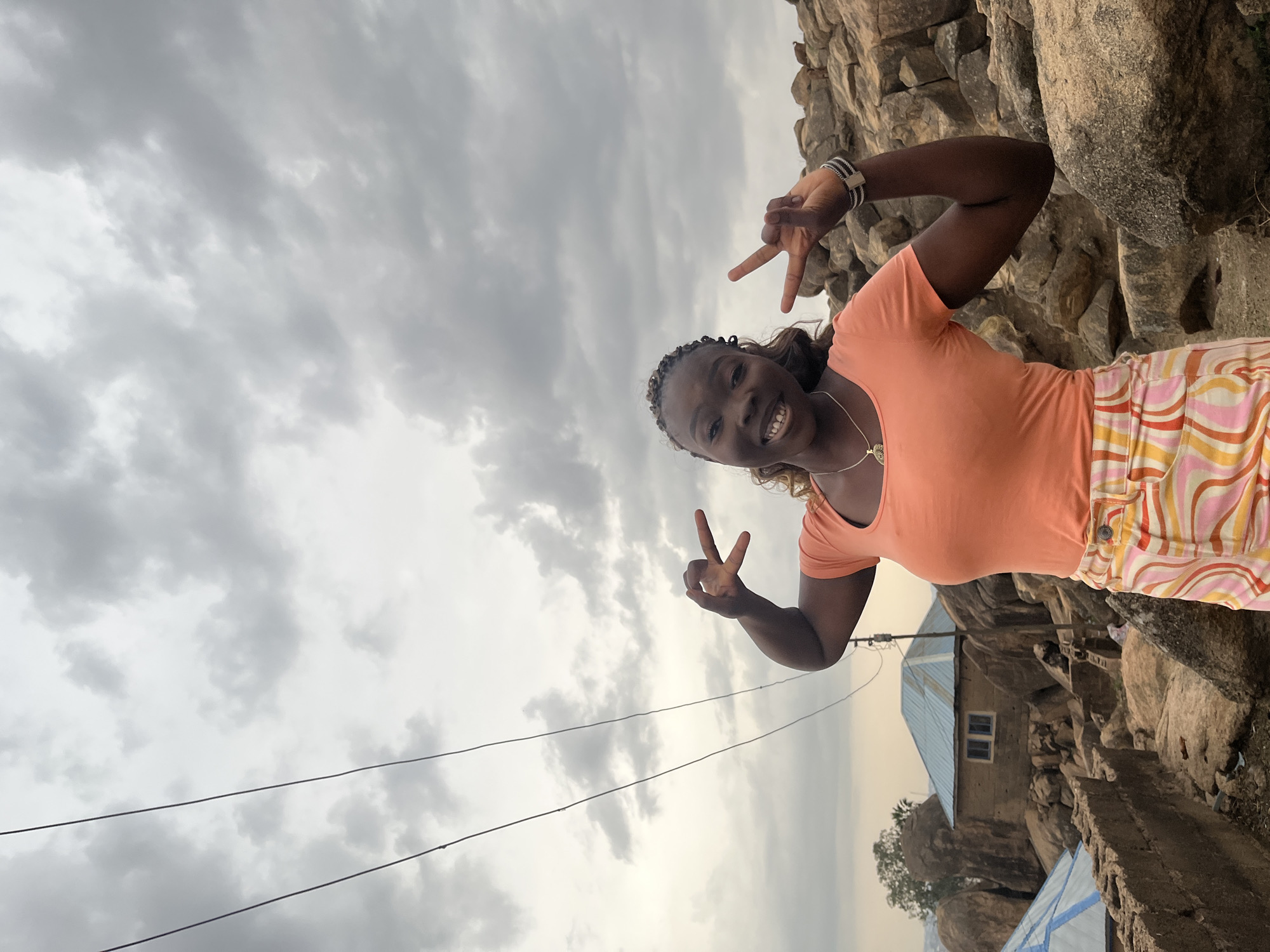 A young woman is posing for a picture outdoors near a large rock formation and a building with a blue roof. The sky is overcast with grey clouds. The woman is wearing an orange shirt and patterned pants. She is smiling and making a peace sign with both hands. In the background, there is a building with a blue metal roof, likely a dwelling or structure within a residential area. There are power lines overhead. The scene is likely taken in or near the city of Jos, Nigeria, judging by the geological formations and vegetation.