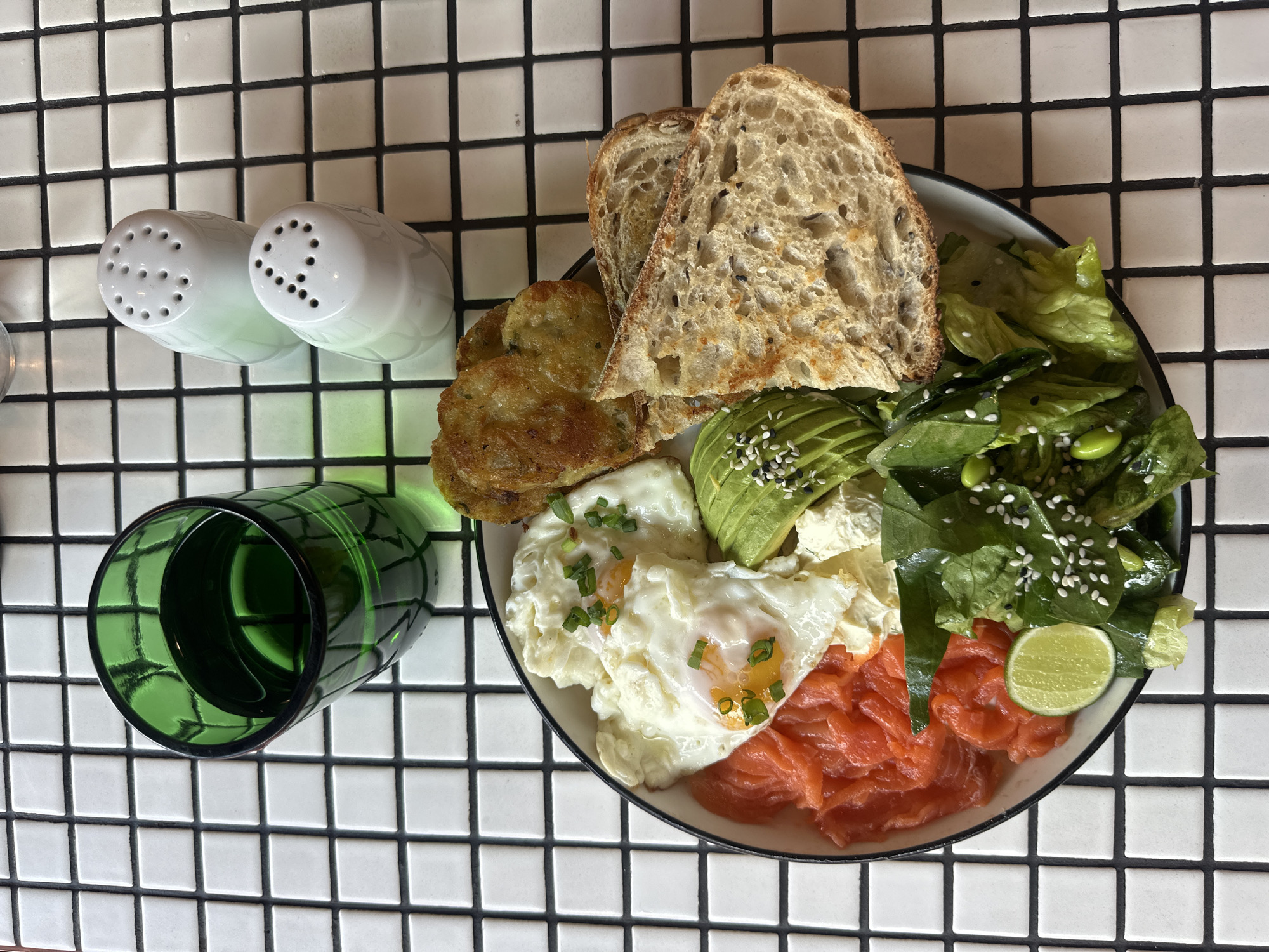 A high-angle, full-shot depicts a plate of food, two salt shakers, and a glass on a checkered surface.
The plate contains two slices of seedy toast, two fried potato cakes, two fried eggs topped with chopped scallions, sliced avocado sprinkled with sesame seeds, a mound of salmon, a pile of leafy green salad with edamame sprinkled with sesame seeds, and a lime wedge.
To the left of the plate are two white salt shakers and a green glass, all sitting on a white and black checkered surface.
This is likely a meal served in North Kuta, Indonesia.