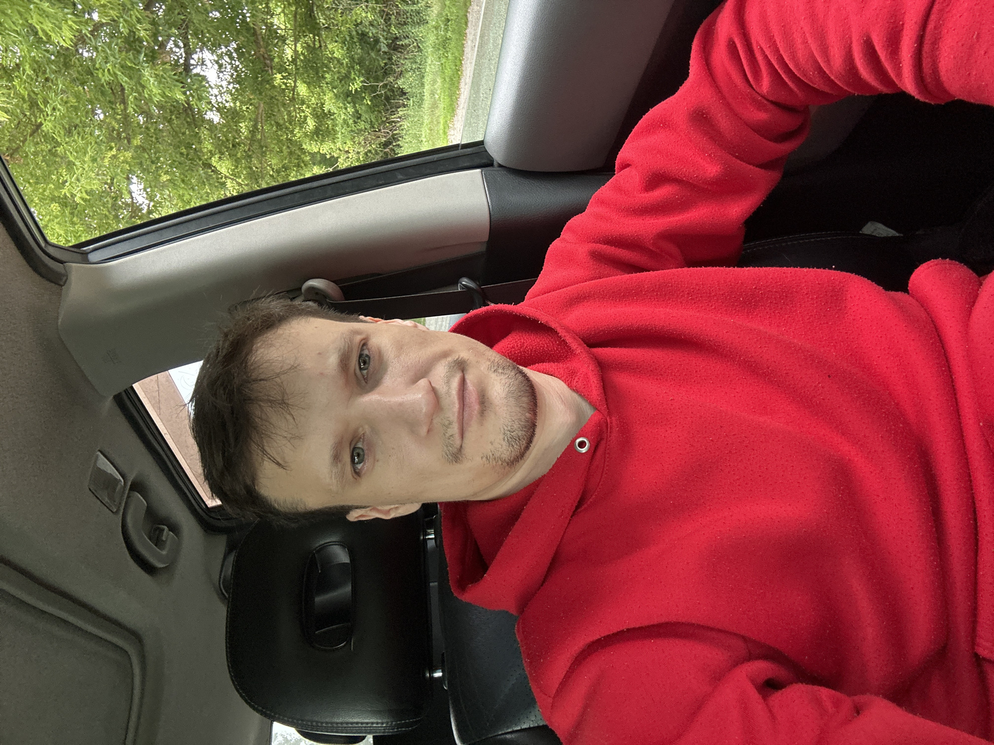 A man is captured from the chest up in a selfie taken from inside a car. He is wearing a red hooded sweatshirt. The man has light skin and short, dark brown hair. He has a neutral expression with his mouth closed and appears to have a light stubble on his chin and upper lip. His eyes are open and looking directly at the camera. Behind him, the black leather interior of the car is visible, including the headrest of the driver's seat and a portion of the car's roof, which has a sunroof. To the right of the man, a car door is visible, and through the open window, lush green trees and foliage are in the background, suggesting an outdoor, possibly rural, setting. The lighting is bright, indicating it is daytime. The location is not specified as Makiv, Ukraine.
