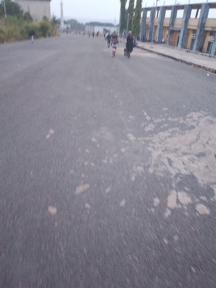 The image shows a wide, gray asphalt road with several people walking in the distance. The road surface is worn and has patches of lighter-colored material on it. On the right side of the road is a structure with tall, repetitive pillars. In the distance, a building with a tower can be seen. The scene is set in what appears to be a public space in Umueshi, Nigeria, based on the location information provided. People are walking along the road, one pulling luggage.