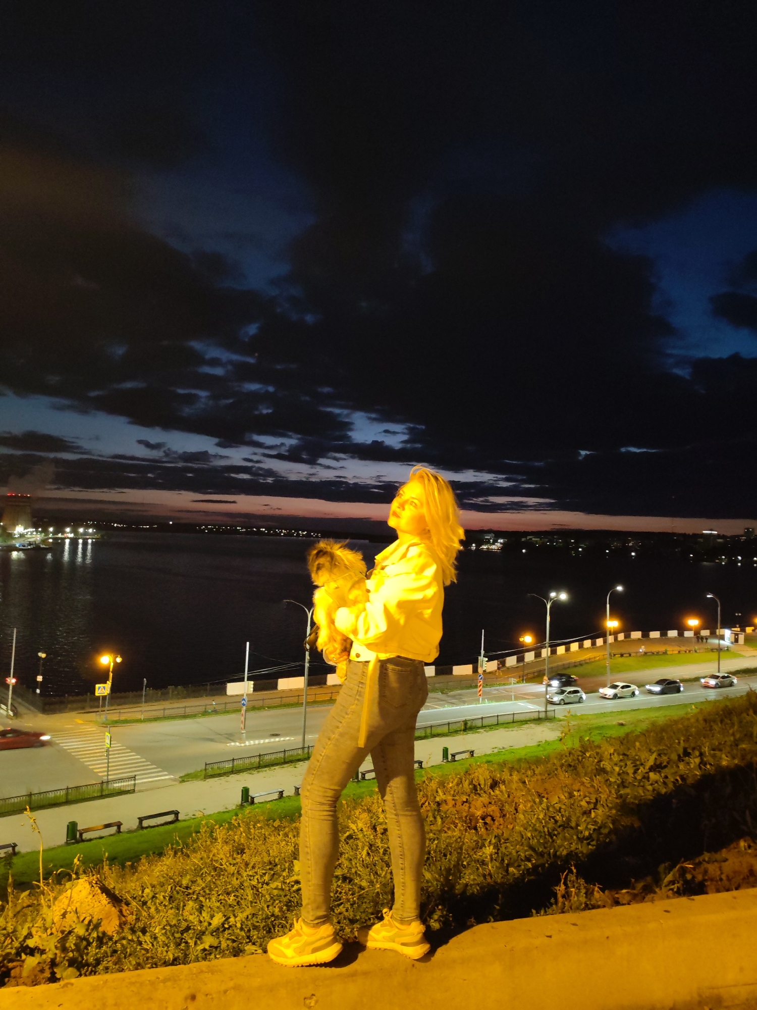 A young woman with blonde hair stands on a grassy embankment at night, holding a small dog. She is wearing a bright yellow jacket and gray jeans. The background shows a body of water, a city skyline with lights, and a road with cars. The sky is dark with some clouds and a hint of twilight colors. The scene appears to be a waterfront promenade in Izhevsk, Russia, possibly near the St. Michael's Cathedral, given the presence of a body of water and a city skyline characteristic of the area.