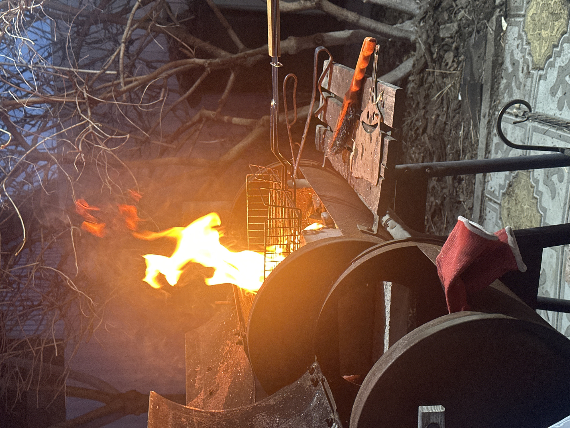 The image depicts an outdoor cooking scene at night or late evening. The central subject is a dark, heavy-duty metal grill or smoker, appearing to be constructed from cylindrical metal barrels or similar components. Bright, lively orange and yellow flames are actively burning within an opening on the left side of the apparatus, emitting smoke or steam.
Above the grill, various cooking utensils are hanging from a wooden support. These include a pair of long metal tongs, a metal grill basket, and a brush with a wooden handle. A notable detail is a metal spatula, also with a wooden handle, which features a distinctive cutout of a smiley face. On the right side of the grill, a red work glove with white trim is casually resting on one of the metal cylinders.
The background on the left is dominated by a dense network of bare tree branches, silhouetted against a dark, possibly twilight or night, sky. This suggests a colder season or a natural, un-leafed tree. On the far right, a vertical surface with an ornate, patterned design, resembling decorative tiles or pavement in shades of yellow-brown and grey-blue, is partially visible.
The activity taking place is clearly outdoor grilling or preparing a fire for cooking. The time of day is dark, illuminated primarily by the firelight, creating a warm glow around the grill. No explicit text is visible, though the smiley face on the spatula is a decorative element. The scene suggests a cozy, possibly rustic, outdoor cooking experience in Krasnoyarsk, Russia.