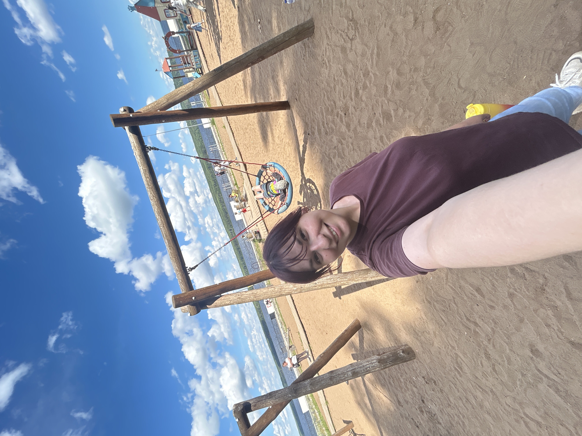 The image shows a young woman taking a selfie at a playground near a body of water, likely the Kama River reservoir in Chaikovsky, Russia. The playground has a wooden swing set, and a child is seen on a net swing in the background. A playground structure with colorful roofs is visible further back, and several people are walking around. The ground is covered in sand. The sky is blue with scattered white clouds. The woman is wearing a maroon t-shirt and blue jeans. A yellow and red water bottle is visible near her.