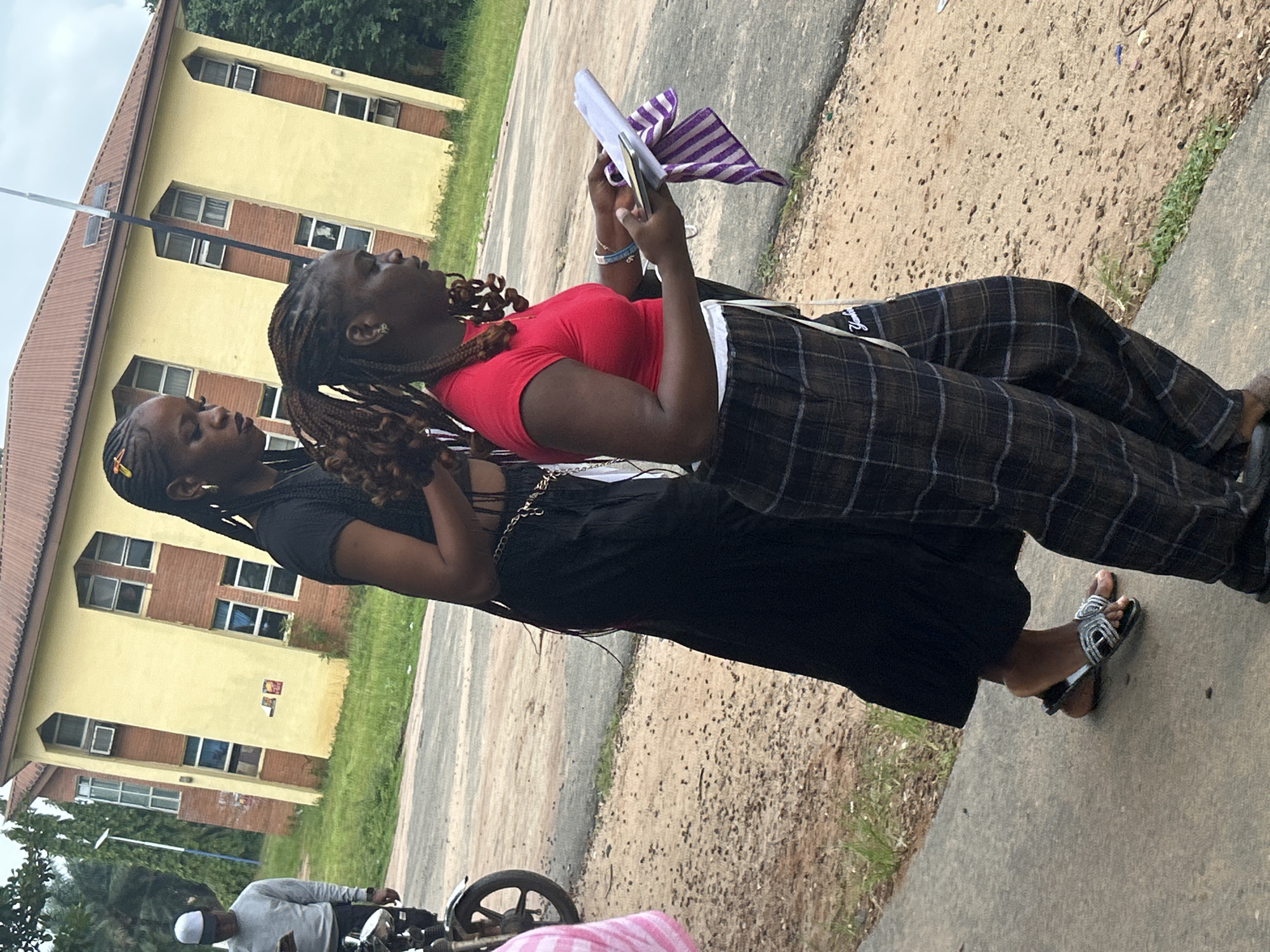 In Ihiagwa, Nigeria, two young women are walking outdoors. The woman in the foreground is wearing a red t-shirt and plaid pants, and she is holding a tablet and a striped cloth. The woman behind her is wearing a black top and a long black skirt. They are walking on a sidewalk in front of a light yellow building with brown brick accents. A motorcycle is parked to the left of the frame, and a man in a grey sweatshirt and white cap is standing near it. The ground is covered in dirt and scattered debris, with patches of grass visible.