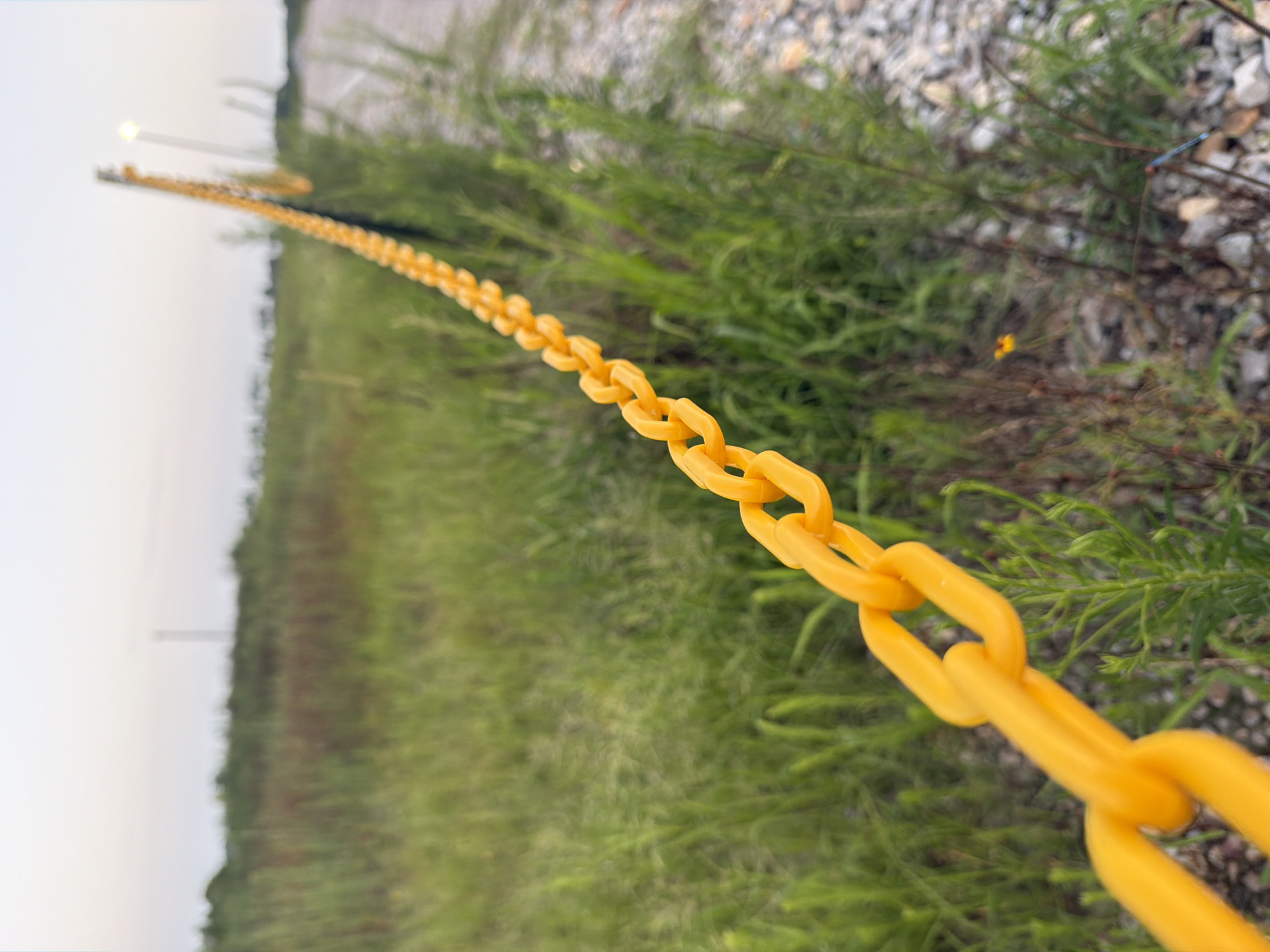 A close-up, low-angle, outdoor shot depicts a bright yellow plastic chain fence stretching diagonally from the lower left corner of the frame towards the upper right. The chain links are large and hollow, forming a repeating pattern against a background of green grass and a gravelly path. In the distance, the chain fence continues, bordered by a light pole with an illuminated lamp. The sky above is a pale, overcast gray, suggesting either dawn or dusk. The environment appears to be a rural or undeveloped area, possibly on the outskirts of Cardwell, United States, with tall weeds and sparse vegetation. There are no people or animals visible in the image.