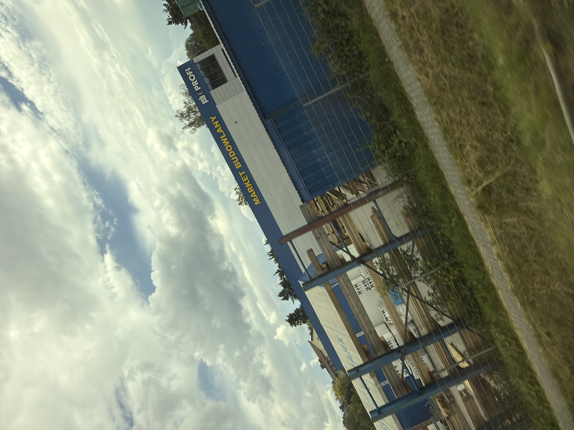 The image shows a low-angle, tilted outdoor shot of a "MARKET BUDOWLANY" (Construction Market) with "psb PROFI" branding. The building is a modern structure with a white and blue facade. The building appears to be a retail store for building materials. In front of the building, there are stacks of construction materials, possibly wood or insulation panels, displayed on metal racks. A metal fence runs along the foreground, separating the construction market from a grassy area and a pathway. The sky above is filled with dramatic clouds. The presence of the "MARKET BUDOWLANY" and "psb PROFI" branding, coupled with the architectural style of the building and surrounding vegetation, strongly suggests this location is in Poland. Based on external knowledge, a PSB PROFI market is located in the city of Wieliczka, Poland, making it highly probable that this image was taken there.