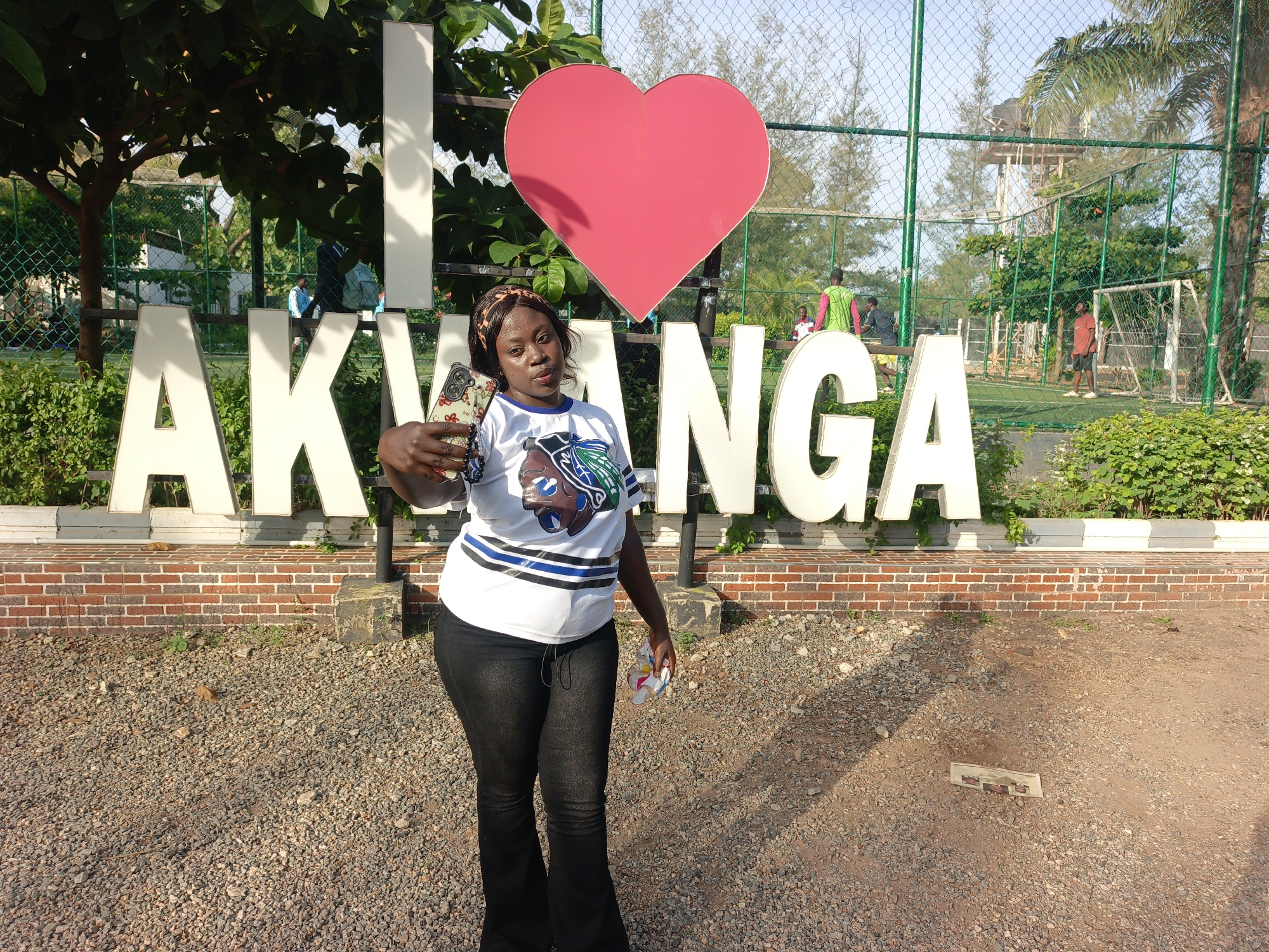 A woman stands in front of the "I ❤️ AKWANGA" sign in Akwanga, Nigeria. She is holding a phone in her left hand, as if taking a selfie. She is wearing a t-shirt with blue and white stripes, black jeans, and a leopard print headband. The sign is white with a red heart. Behind the sign, there is a fenced-in soccer field with players. The ground is covered in gravel.