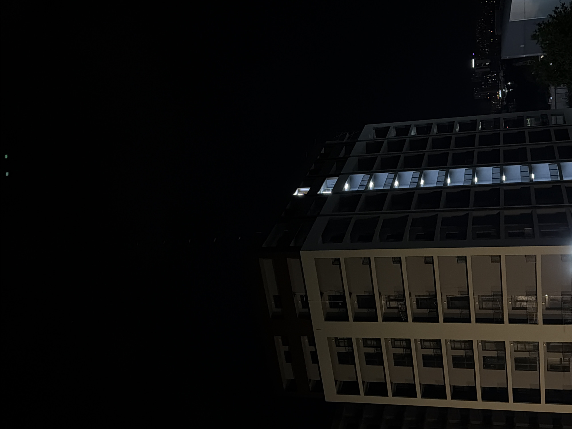 The image depicts a nighttime view of a modern apartment building in Tbilisi, Georgia. The building is tall and appears to be constructed with concrete and glass, featuring multiple balconies and illuminated windows. The dark sky contrasts with the lit portions of the building, creating a dramatic effect. In the background, other buildings and city lights are visible, suggesting an urban setting. The overall impression is one of a quiet, residential area at night, with the architecture of the building being a prominent feature of the scene. There are no people visible in the image.