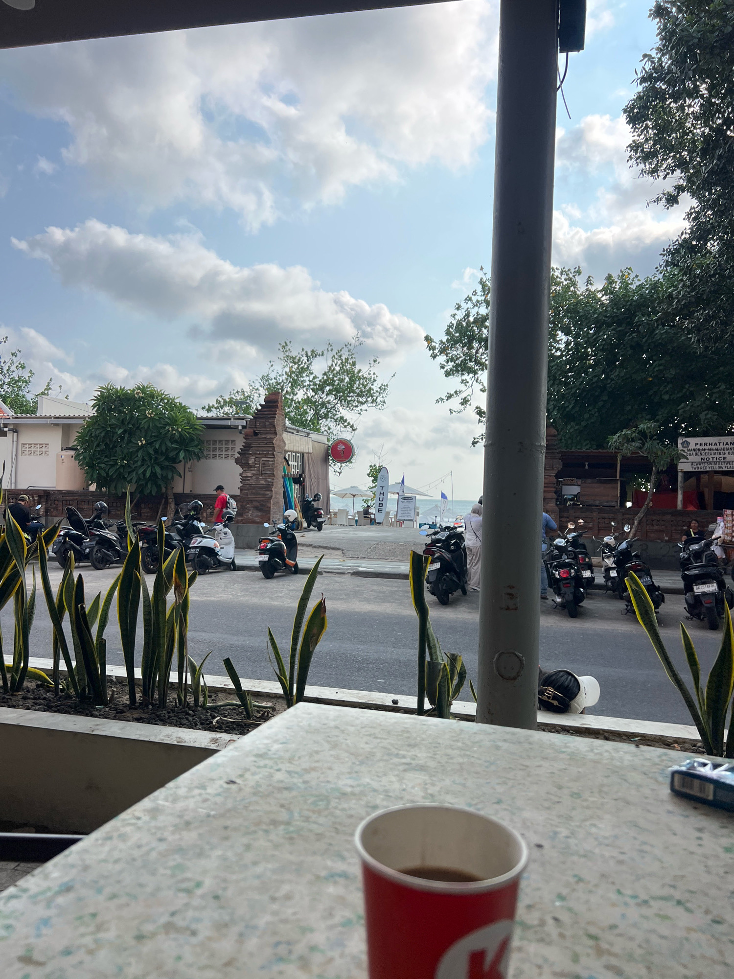 The image captures a street scene in Kuta, Indonesia, likely near the beach. In the foreground, a speckled countertop is visible, with a red paper cup filled with a dark liquid (likely coffee) and a partially open pack of cigarettes or a small electronic device on the right. A large, grey cylindrical pillar dominates the right side of the frame, obscuring part of the background. Beyond the pillar, a road is lined with parked motorbikes, mostly scooters. Several people are seen near the motorbikes and walking on the sidewalk.
To the left of the pillar, a planter box with tall, green, sword-like plants is present. Behind this, a white building with visible brickwork and a partially ruined wall stands. Further back, trees and vegetation can be seen. In the distance, a glimpse of the ocean and beach is visible, with some umbrellas and beach chairs suggesting a tourist area. The sky is partly cloudy, with patches of blue. A sign with Indonesian text and the word "NOTICE" is affixed to a wall on the right, indicating a public area with rules or information. The overall atmosphere suggests a sunny day in a bustling, coastal tourist town.