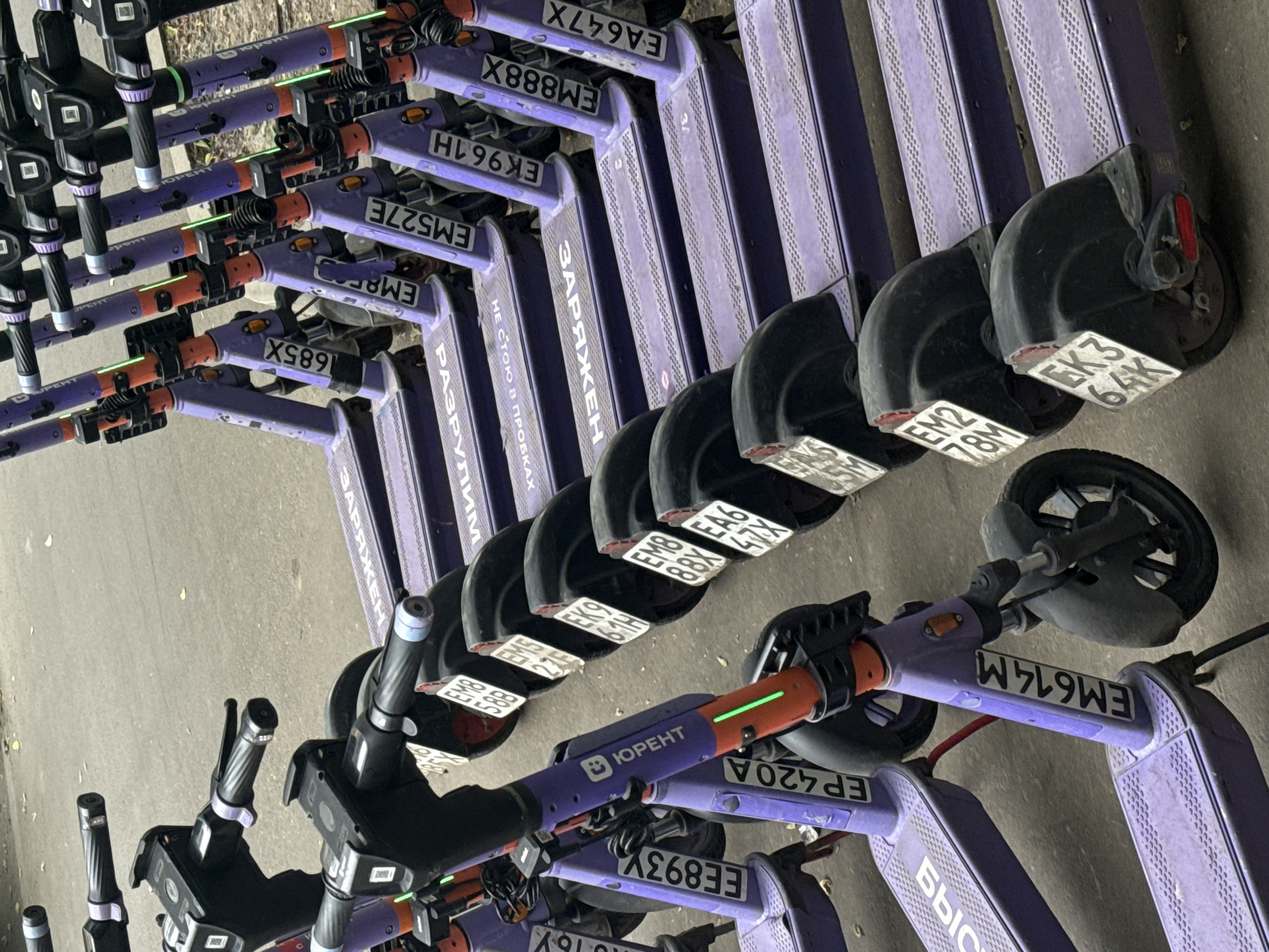 The image shows a row of parked purple electric scooters on a paved surface, likely a sidewalk or street in Moscow, Russia. The scooters are lined up facing towards the right of the frame. Each scooter has a black handlebar with a digital display and grips, a purple stem with an orange accent and a green LED light, and a purple deck. Some scooters have license plates with alphanumeric codes and the "ЮRENT" logo. The ground has a textured pattern on the decks of the scooters, and the background shows a blurry asphalt surface. The scene is static, with no people or any discernible action taking place. The overall impression is of a rental service preparing for use.