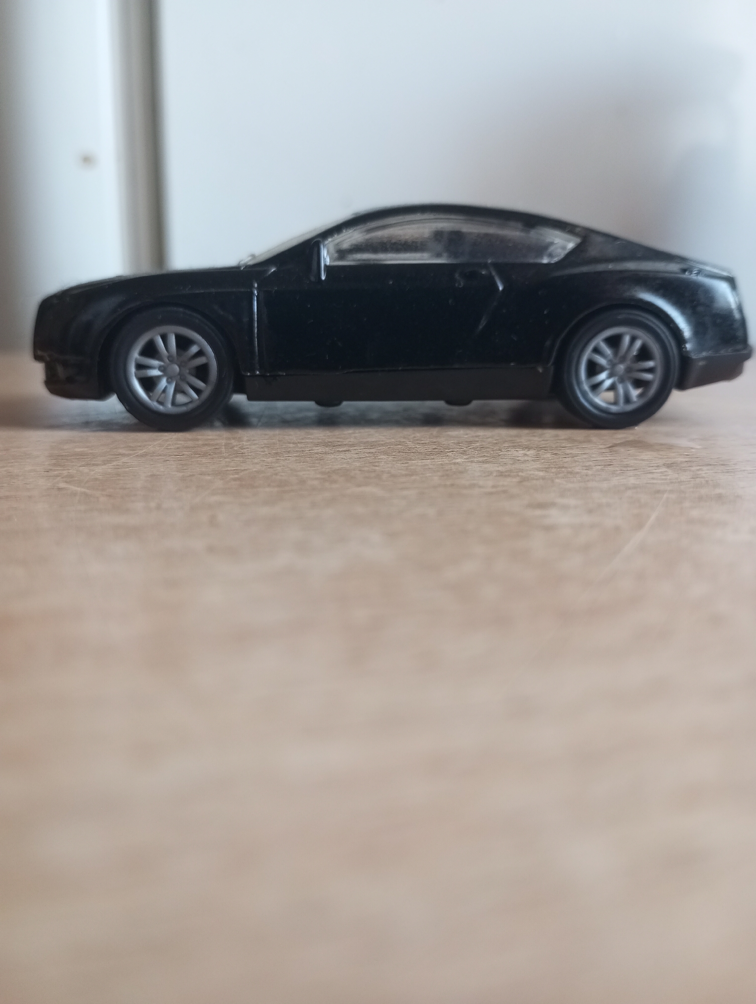 A black toy car is positioned on a wooden surface. The car is a coupe with a sleek design, and its wheels are visible. The background is blurred, but it appears to be an indoor setting with a light-colored wall. The image is taken from a side profile of the car, focusing on its details. There are no people or any other objects in the frame besides the car and the surface it rests on. The lighting suggests it might be natural light coming from a window, with some shadows cast on the surface.