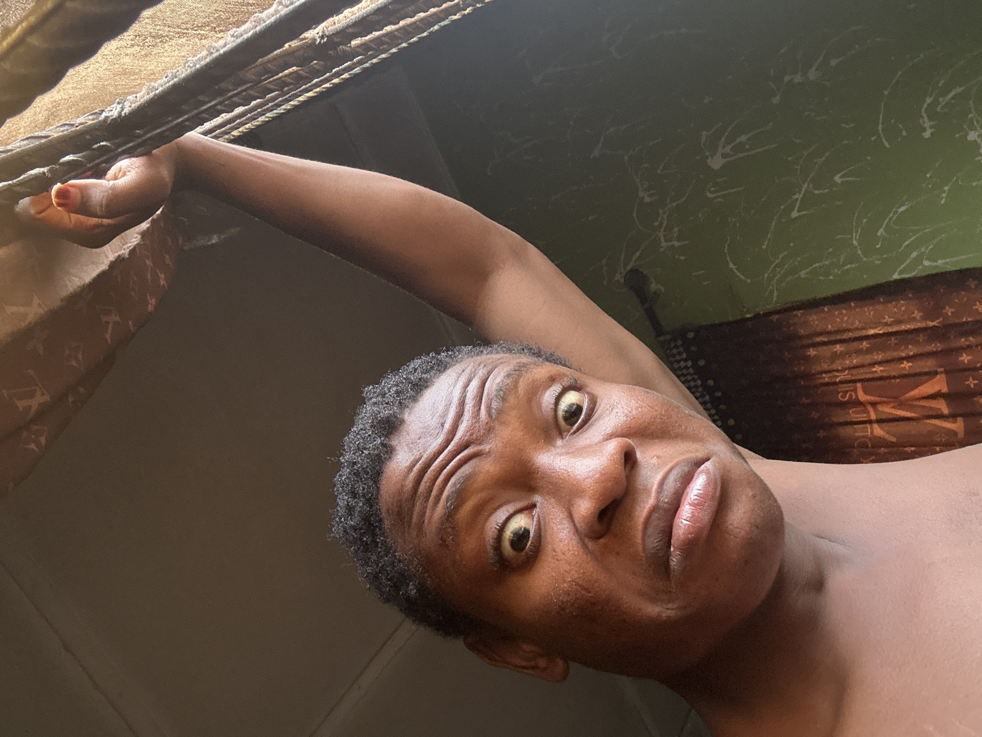 A close-up, low-angle shot depicts a person of African descent with short, dark, tightly curled hair. The individual has noticeable lines on their forehead and a somewhat perplexed expression, their eyes wide and lips pursed. They are grasping onto what appears to be a metal bar or frame, possibly the top of a window. The arm holding the bar is extended, showing skin tone consistent with the person's face.
In the background, a green wall with a white splatter pattern is visible. Below the wall is a brown and tan striped pattern. The setting is indoors, but the specific location is indeterminable.