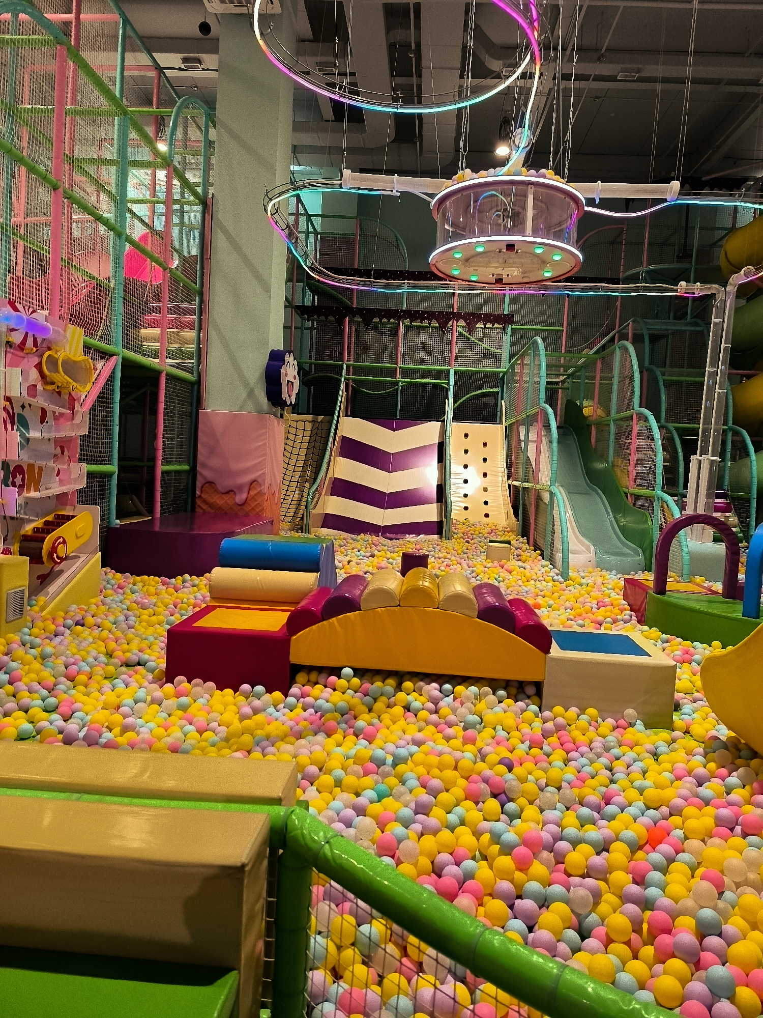 An indoor children's play area is depicted, filled with a vast quantity of colorful balls in shades of yellow, pink, blue, and purple. The play area features various soft play structures, including padded seating elements and a ball pit. In the background, a multi-level climbing structure with netting and slides is visible. Neon lighting accents the ceiling and parts of the play structure, creating a vibrant atmosphere. A cylindrical suspended ride with lights is also present. The image does not provide information to ascertain its location in the city of Qarağandı city administration, Kazakhstan. There are no people visible in the image.