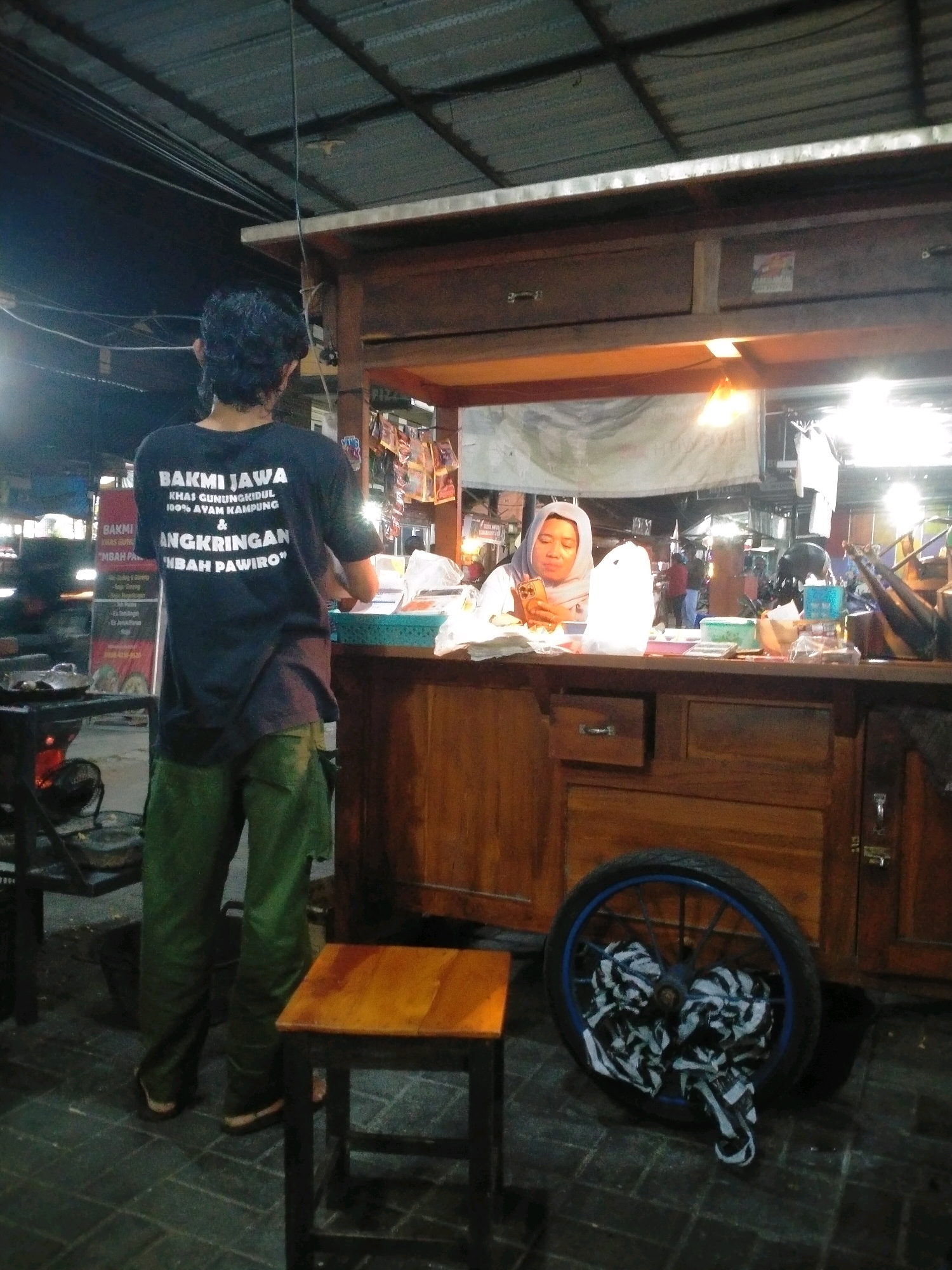 A man wearing a dark t-shirt with white text and green pants stands with his back to the camera at a food stall in Kasihan, Indonesia. The t-shirt text appears to be in Indonesian, with "BAKMI JAWA" prominently displayed. The stall, constructed of dark wood, has a counter where a woman wearing a light-colored hijab is seated, looking at her phone. She is holding a white paper bag and there are various items on the counter, including plastic containers, a calculator, and a stack of white paper. Behind the counter, merchandise is displayed on shelves. A small wooden stool is positioned in front of the stall, and a spoked wheel with black and white fabric tied to it rests on the tiled ground to the right. A cooking area with a wok is visible to the left of the man, with flames visible beneath it. The overall lighting suggests it is evening or nighttime, with artificial lights illuminating the scene.