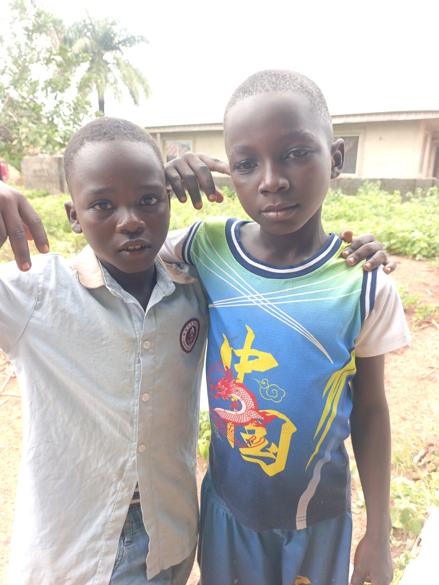 Two young boys are standing side-by-side, looking at the camera, in what appears to be the exterior of a building. The boy on the left is wearing a light blue collared shirt with buttons and a circular emblem on the left chest. He has his left arm extended, with his hand resting on his thigh and his right arm over the shoulder of the boy next to him. The boy on the right is wearing a blue, green, and white t-shirt with a dragon design and yellow accents. His left arm is resting on the boy on the left's shoulder. Both boys have short hair. The background consists of greenery, a building with windows, and an overcast sky, suggesting an outdoor setting in Akwanga, Nigeria.