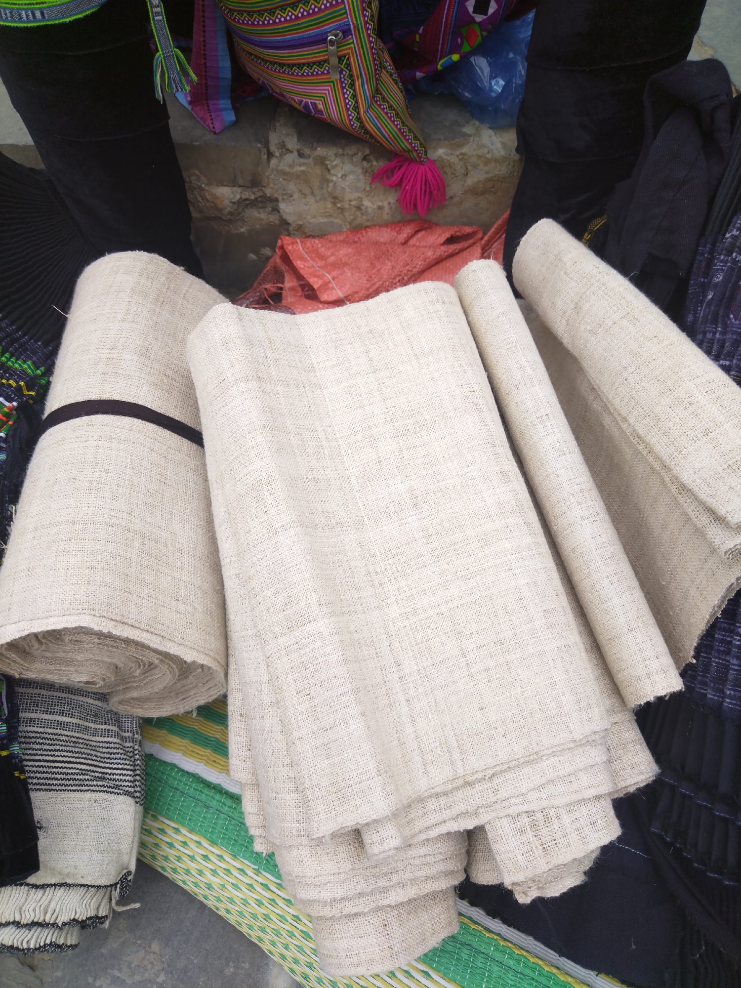 The image depicts a collection of various fabric rolls and textiles. The most prominent features are several rolls of off-white, woven fabric, likely made of linen or a similar natural fiber. One roll is secured with a dark brown band. The fabric rolls appear to be stacked or arranged casually. In the background, there are glimpses of colorful textiles and a decorative bag with pink tassels, suggesting the items might be part of a market or display selling textiles. Also visible are other fabric pieces with intricate patterns and dark colors. The location is unspecified, but the variety of fabrics and textiles suggests a vibrant trade or craft setting.