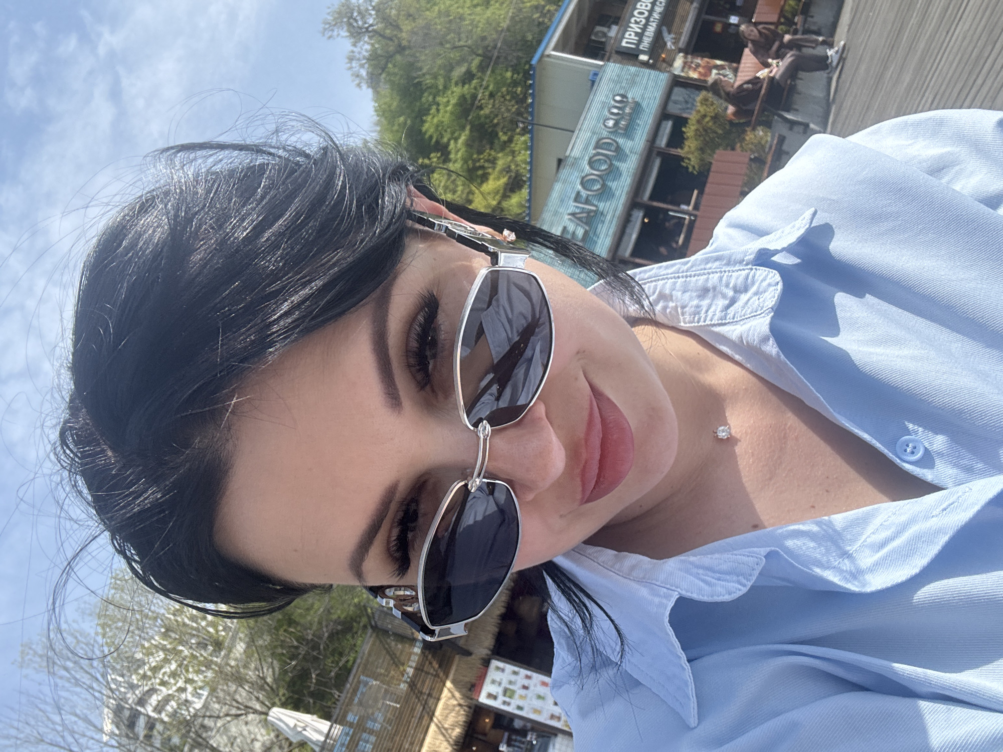 A woman with dark hair and sunglasses is taking a selfie. She is wearing a light blue button-down shirt. A building with the word "SEAFOOD" is visible in the background. Text in Cyrillic script is above the word "SEAFOOD." Based on the Cyrillic script and the restaurant name, it is likely that the image was taken in Vladivostok, Russia.
