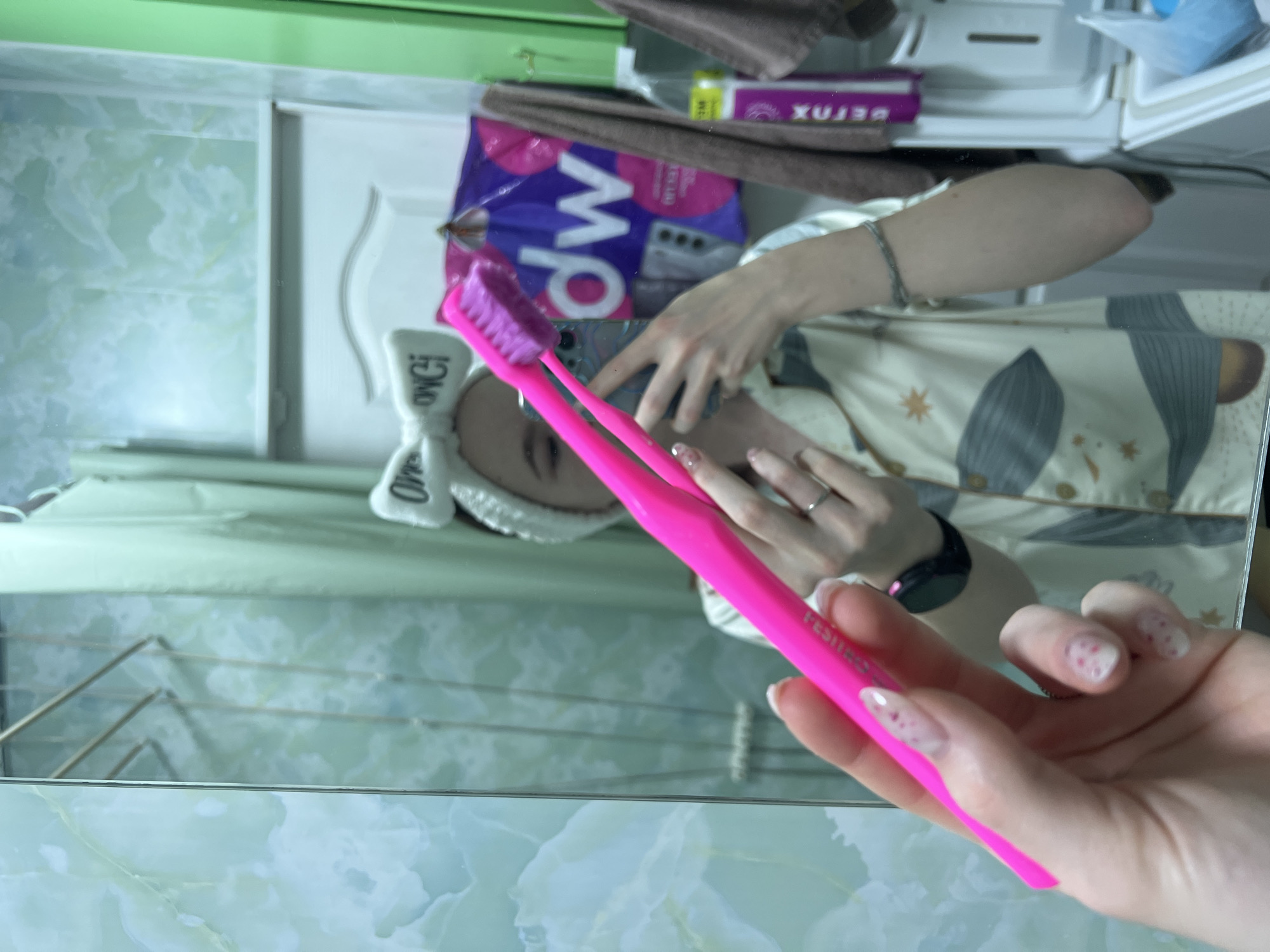 A person is holding a pink toothbrush in their right hand, facing a mirror. The person has a white headband on. The mirror reflects the person's face, hands, and a shelf with various items, including a package labeled "dw" and a box labeled "DETOX." The person is wearing a patterned robe, and a smartwatch is visible on their left wrist. The background includes a green wall with a patterned design. The image appears to have been taken indoors.
The location is not discernible from the image.