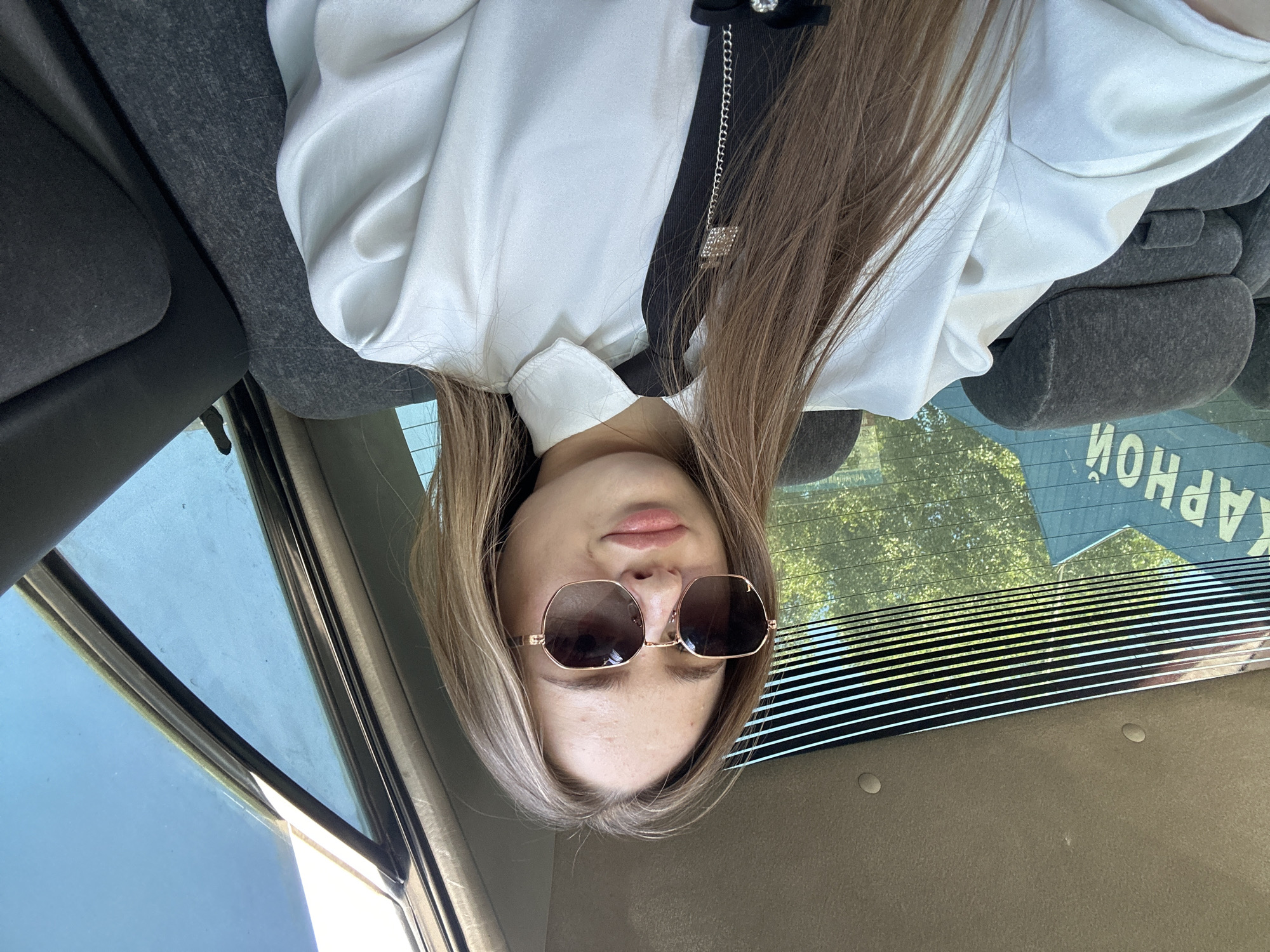 A young woman with blonde hair and sunglasses sits in the back seat of a car, looking at the camera. She is wearing a white collared shirt and a black tie. The car's interior is visible, including the seats and the rear window. Through the window, trees and blue sky are visible, as well as some lettering on the outside of the car. The image appears to be a selfie taken by the woman.