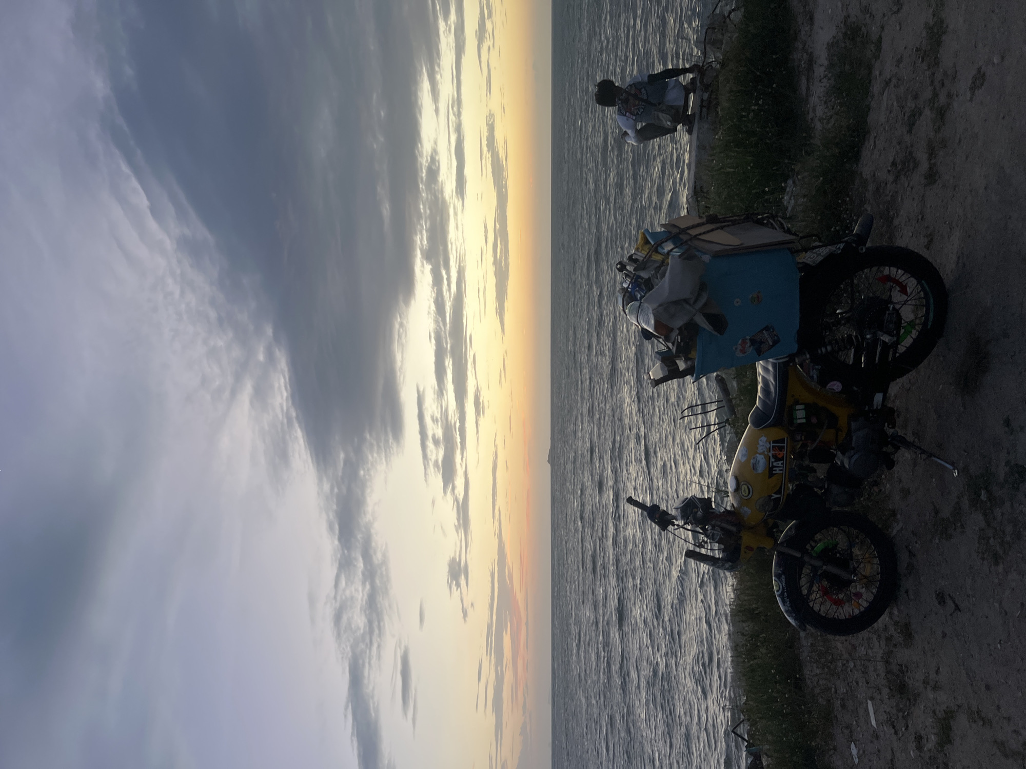 In the city of Ko Pha-ngan District, Thailand, at dusk, a yellow motorcycle is parked on a dirt path overlooking a body of water. The motorcycle is laden with various items in a light blue bin attached to the rear. To the right, a person wearing a white shirt and dark pants is crouched down, facing the water. The sky above is filled with clouds, with the horizon displaying a gradient of orange and yellow hues from the setting sun. The water's surface is rippled, suggesting a gentle breeze. Green vegetation grows along the edge of the embankment.