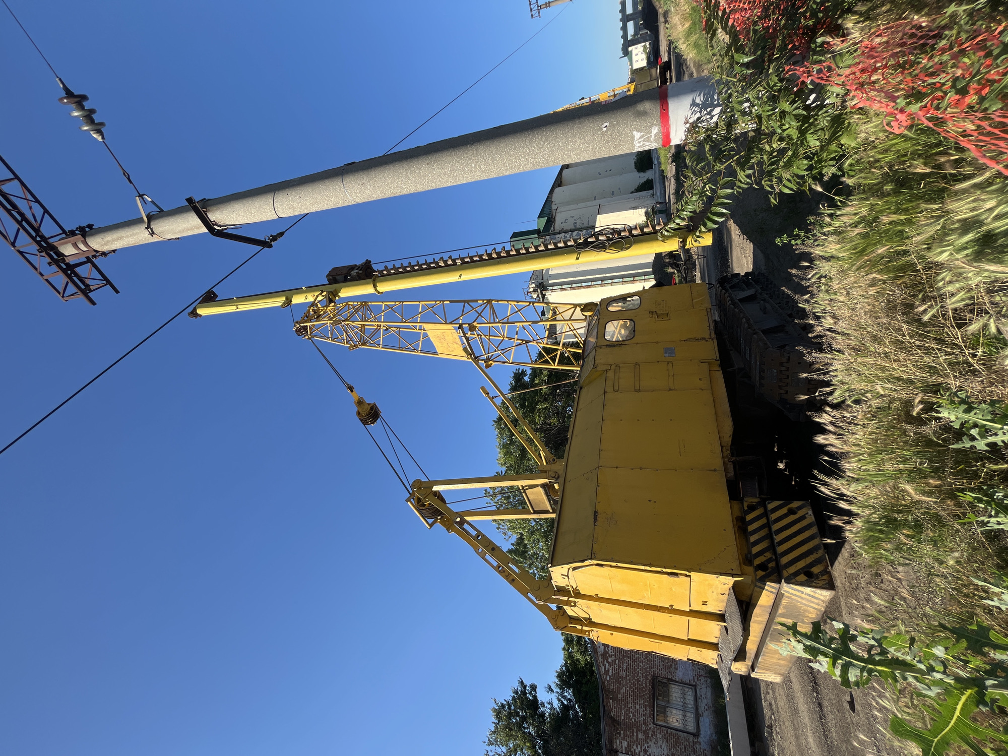 A yellow tracked crane is parked on a dirt lot next to a concrete utility pole and a brick building. The crane has a long, yellow boom extending upwards. There are electrical wires running across the sky. The area appears to be an industrial or construction zone. The sky is clear and blue. The vegetation in the foreground is tall grass and some red foliage. The image is taken from a low angle.