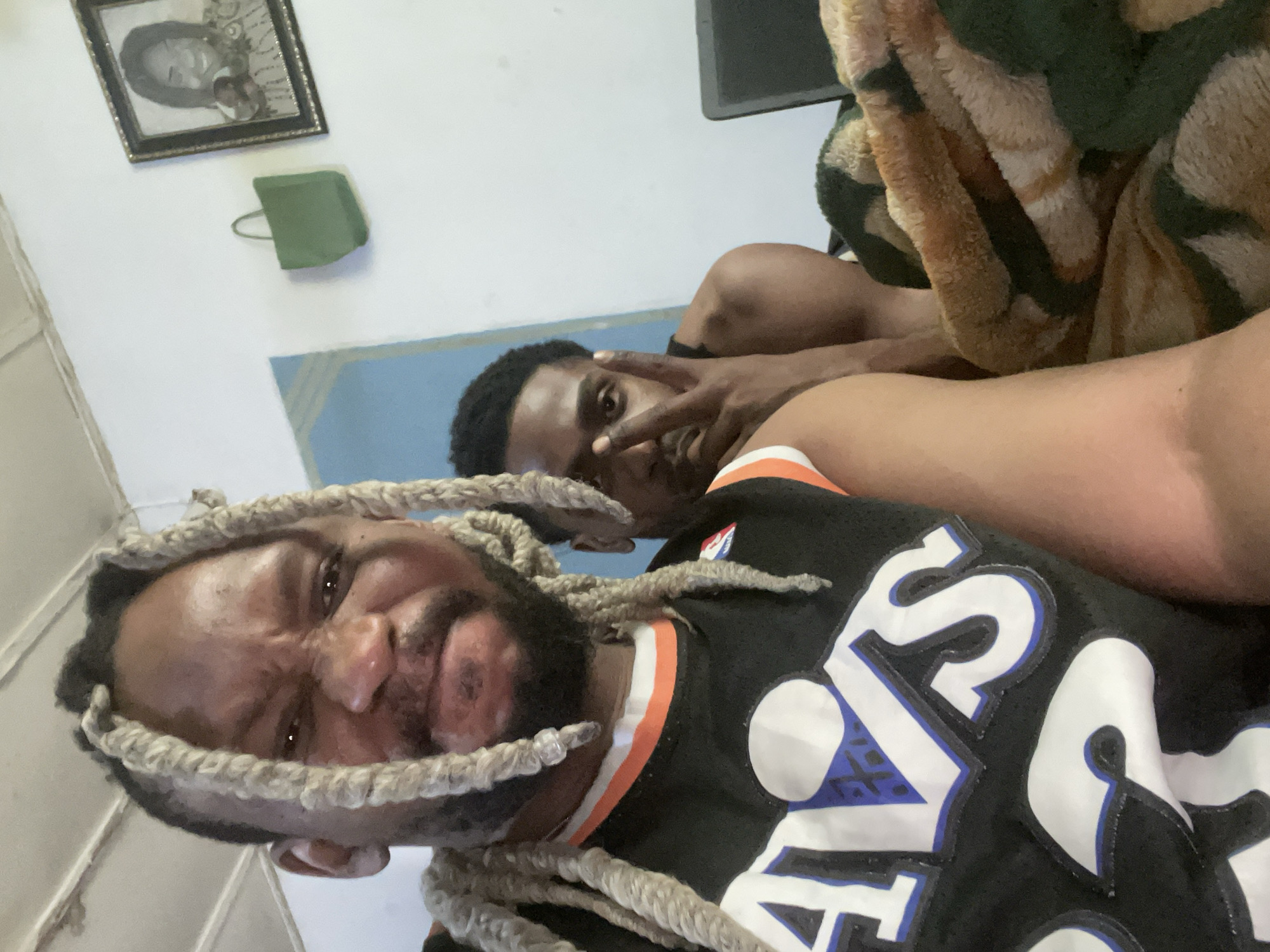The image shows two men lying down in what appears to be an indoor location. The man in the foreground has blonde braids, a dark beard, and is wearing a black sports jersey with orange and white details. His facial expression is strained. The man in the background is darker-skinned with short black hair. His hand is raised near his face in a gesture. The background includes a white wall, a framed picture, a small green object, and a blue section on the wall. A laptop is visible in the upper background along with a patterned blanket. There are no clues within the image to verify the location is in Jos, Nigeria.
