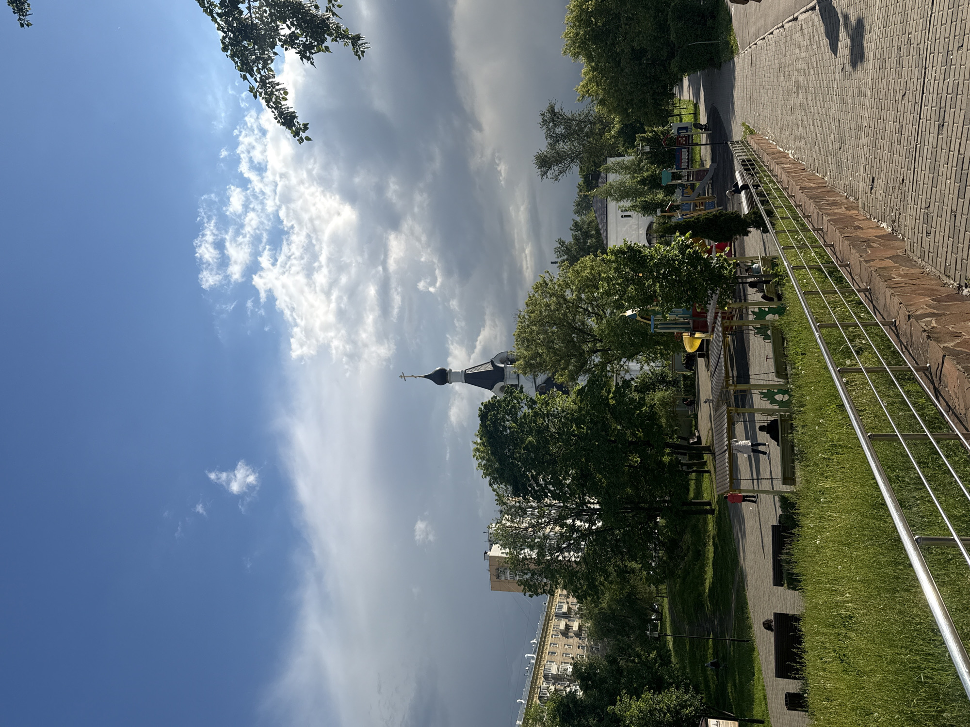Here's a detailed description of the image:
The image shows a park setting with a view of a church, possibly located in a residential area. The sky is partially covered with clouds, creating a dynamic light and shadow effect.
In the foreground, a brick walkway runs parallel to a grassy area. A metal railing separates the walkway from the grass. Several benches are present, indicating a space for people to rest and relax. People are seen walking along the path.
Beyond the park area, a church with a distinctive spire is visible, partially obscured by trees. In the background, apartment buildings are visible. The photo appears to be taken on a sunny day, with a contrast between the bright sky and the darker shaded areas.