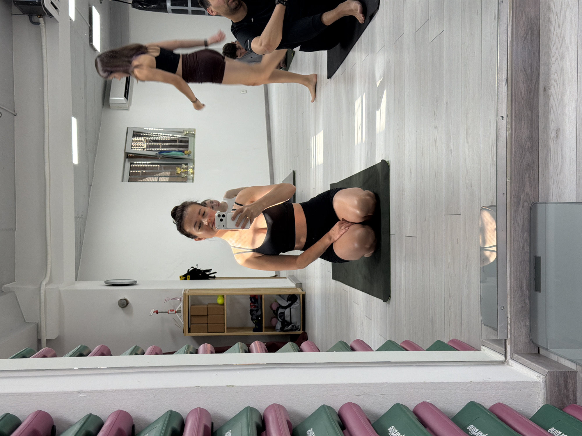 A full shot captures the reflection in a large mirror of a yoga studio in Budva, Montenegro. The studio is illuminated by white overhead lights. The mirror frame is white and the foreground of the reflection shows rows of yoga mats stacked in alternating green and pink.
In the reflection, a woman in a black sports bra and black shorts is kneeling on a green yoga mat, taking a selfie with her smartphone. Behind her, other individuals are participating in a yoga class. A woman in a black top and brown shorts is in motion as if she's running or moving quickly. A man in black athletic wear is also practicing yoga on a black mat. The studio has light wood floors. In the background, there's a window and a shelf containing yoga blocks and other equipment.