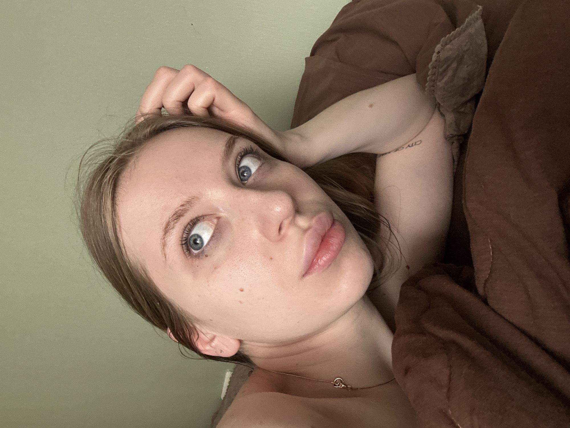 A fair-skinned woman with blonde hair and blue eyes is reclining in bed, partially covered by a brown duvet. She appears to be looking directly at the viewer. Her left hand is resting on her forehead. She is wearing a delicate gold necklace. A tattoo is visible on her upper right arm. The background is a pale green wall. The location is not evident in the image and cannot be confirmed as Chaikovsky, Russia.