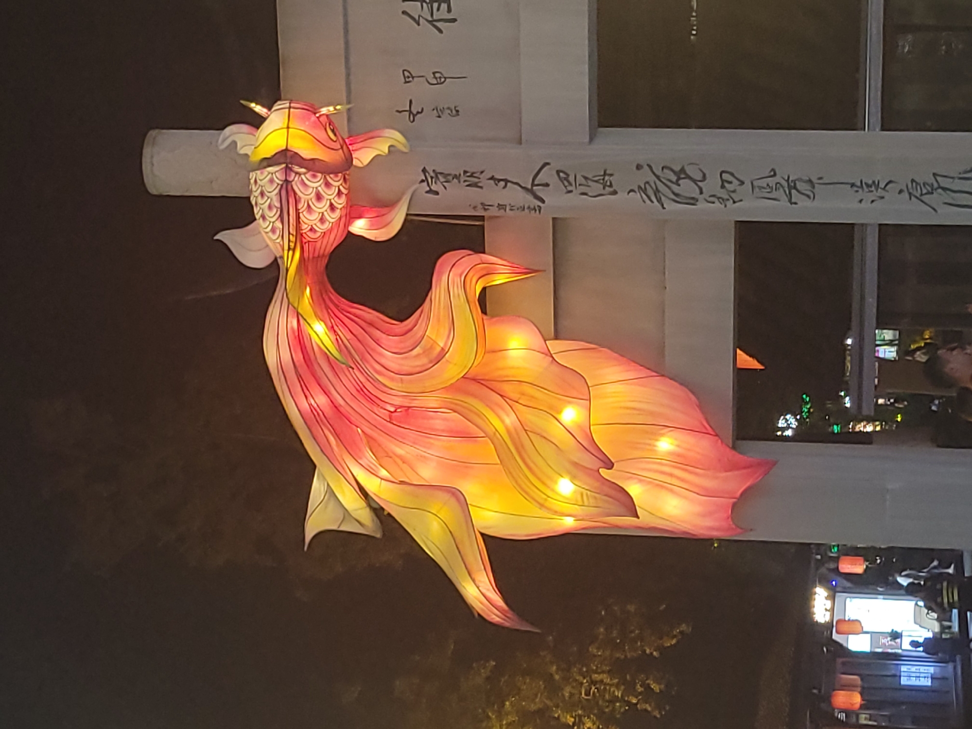 An illuminated, ornate lantern in the shape of a golden-red fish hangs from a white pillar at night in Wuhou District, China. The fish lantern is detailed with scales and fins, and appears to be part of a celebratory decoration. The pillar is inscribed with vertical black Chinese calligraphy. In the background, obscured by darkness and the illumination of the lantern, are faint structures that appear to be buildings with more red lanterns hanging, suggesting a lively night market or festival. There are also silhouetted figures of people in the distance, some walking and others standing. The environment is outdoor, under a dark night sky.