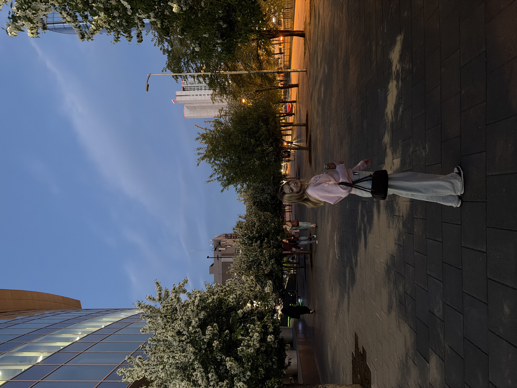 A girl stands on a stone paved pedestrian walkway in the city of Yekaterinburg, Russia, under trees that line the path. Behind her are other pedestrians, and beyond them buildings. Tall structures are visible in the distance. The picture was taken at dusk or twilight, as evidenced by the lighting in the sky.