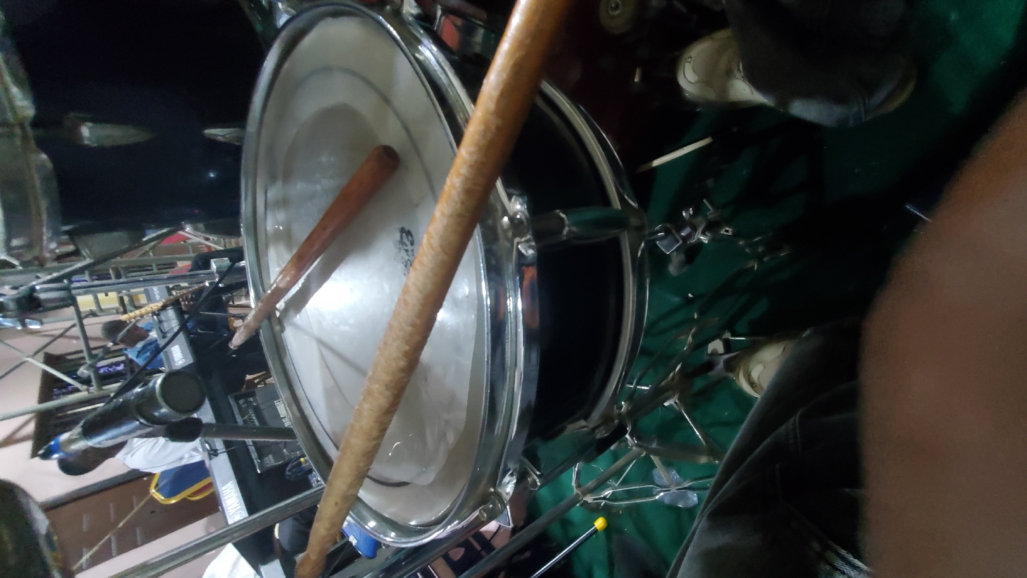 The image depicts a close-up view of a drum set, with two drumsticks resting on the snare drum. In the background, two men are visible. One man, wearing a white shirt, is seated behind a Yamaha keyboard. The other man, in a blue shirt, is playing a keyboard instrument. The setting appears to be indoors, possibly a performance space, with stage lighting and equipment visible. The overall ambiance suggests a musical event or rehearsal. The specific location in Jalingo, Nigeria, is not discernible from the image itself, as there are no geographical markers or distinctive architectural features that could pinpoint the city.