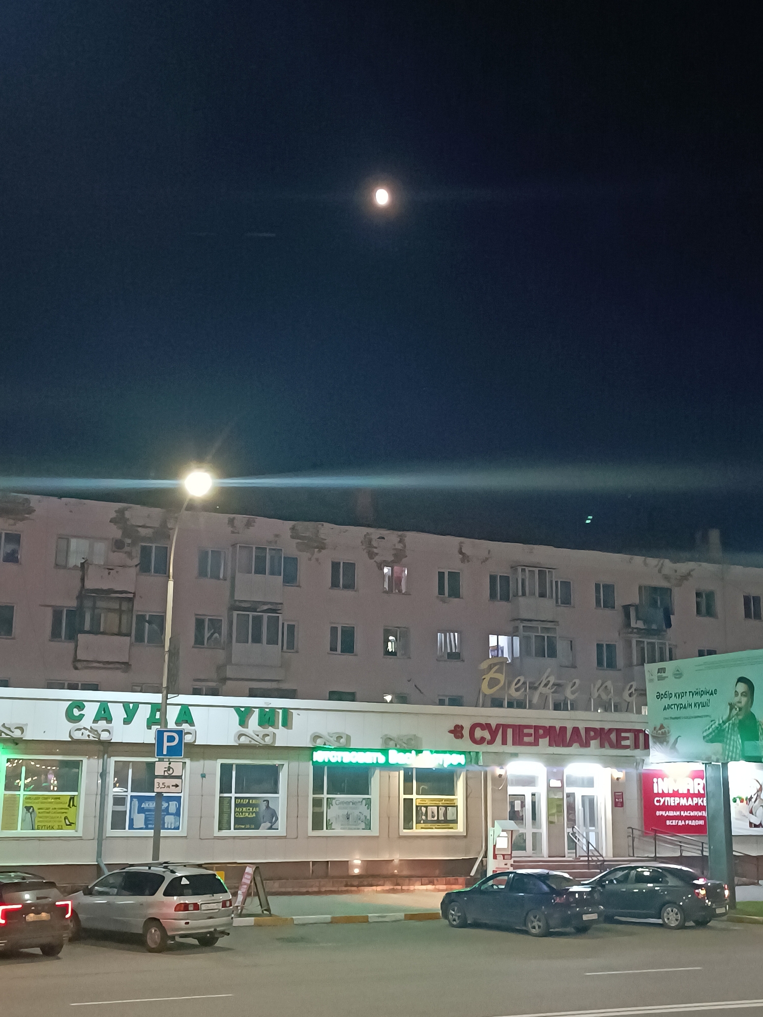 The image captures a nighttime street scene in Kokshetau, Kazakhstan. The sky is dark with a bright full moon. A street light illuminates a building with several floors and multiple windows. The lower level of the building houses a supermarket. The signs above the shop fronts include the Cyrillic characters "САУДА YUI" and "СУПЕРМАРКЕТІ", as well as "Bepepe". Parking signage indicates that parking is available. Several parked cars are visible in the foreground. Another sign advertises a male clothing store. The building's exterior shows some signs of weathering.