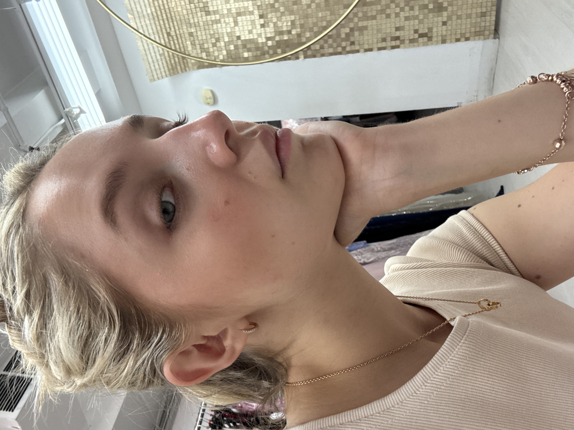 A young blonde woman is pictured in a close-up, side profile shot, resting her chin on her hand. She is wearing a cream-colored ribbed t-shirt and a delicate gold necklace with a pendant. Her hair is pulled back in a messy bun, and she has subtle makeup on, including blush and mascara. A small hoop earring with what appear to be diamonds is visible in her left ear. The background is somewhat out of focus but suggests an indoor setting with racks of clothing and bright overhead lighting. The overall impression is one of a casual, possibly a fitting room or boutique environment. There is no identifiable information in the image to suggest a specific location in New York City, United States of America.