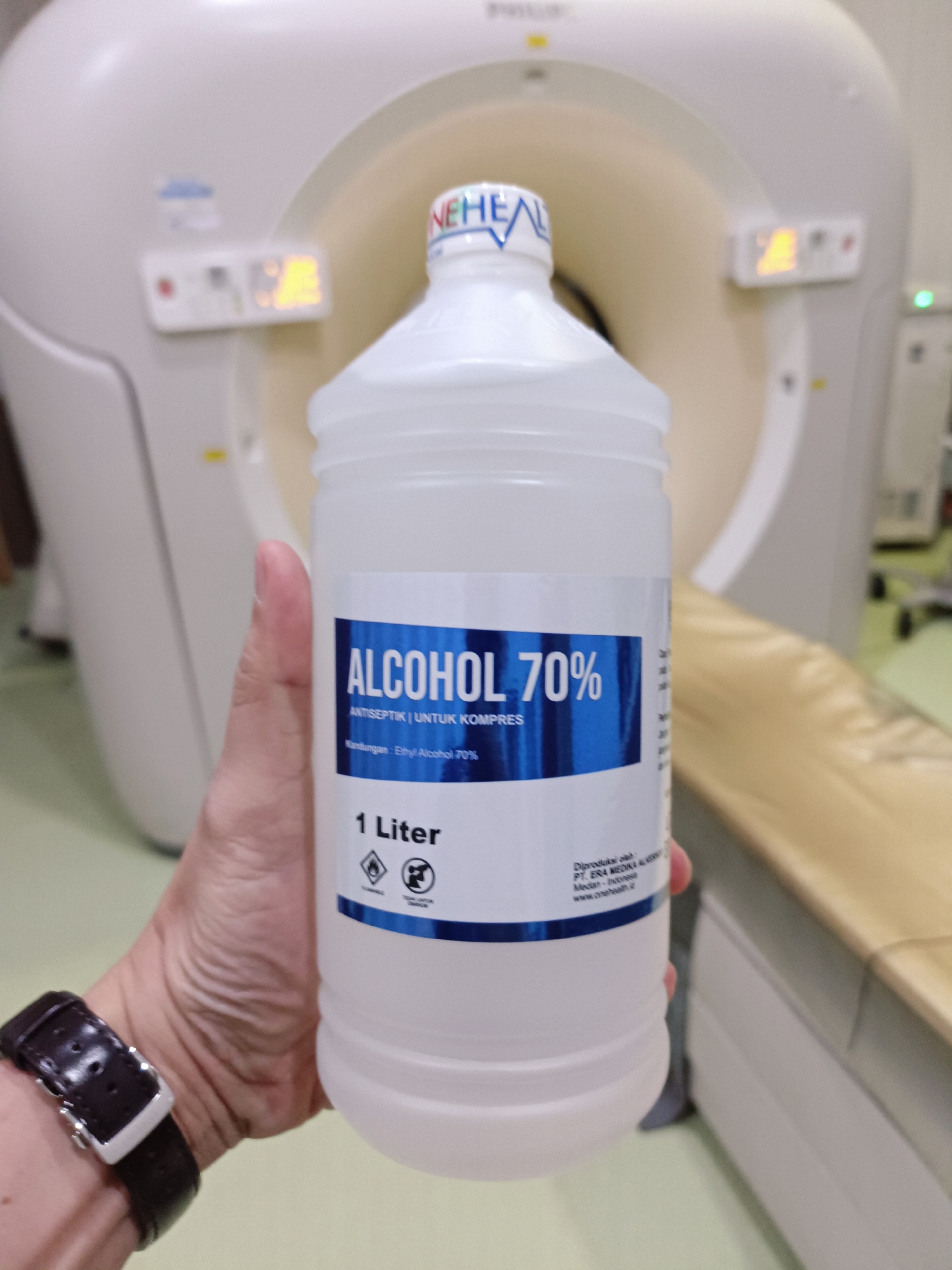 A person's hand, wearing a dark leather watch with a silver buckle, holds a clear plastic bottle of 70% alcohol antiseptic in front of a large, white medical imaging machine, likely a CT scanner. The bottle has a blue and white label that reads "ALCOHOL 70%" and "1 Liter". The machine in the background appears to be in an indoor, sterile environment, suggested by the presence of medical equipment and the lighting.