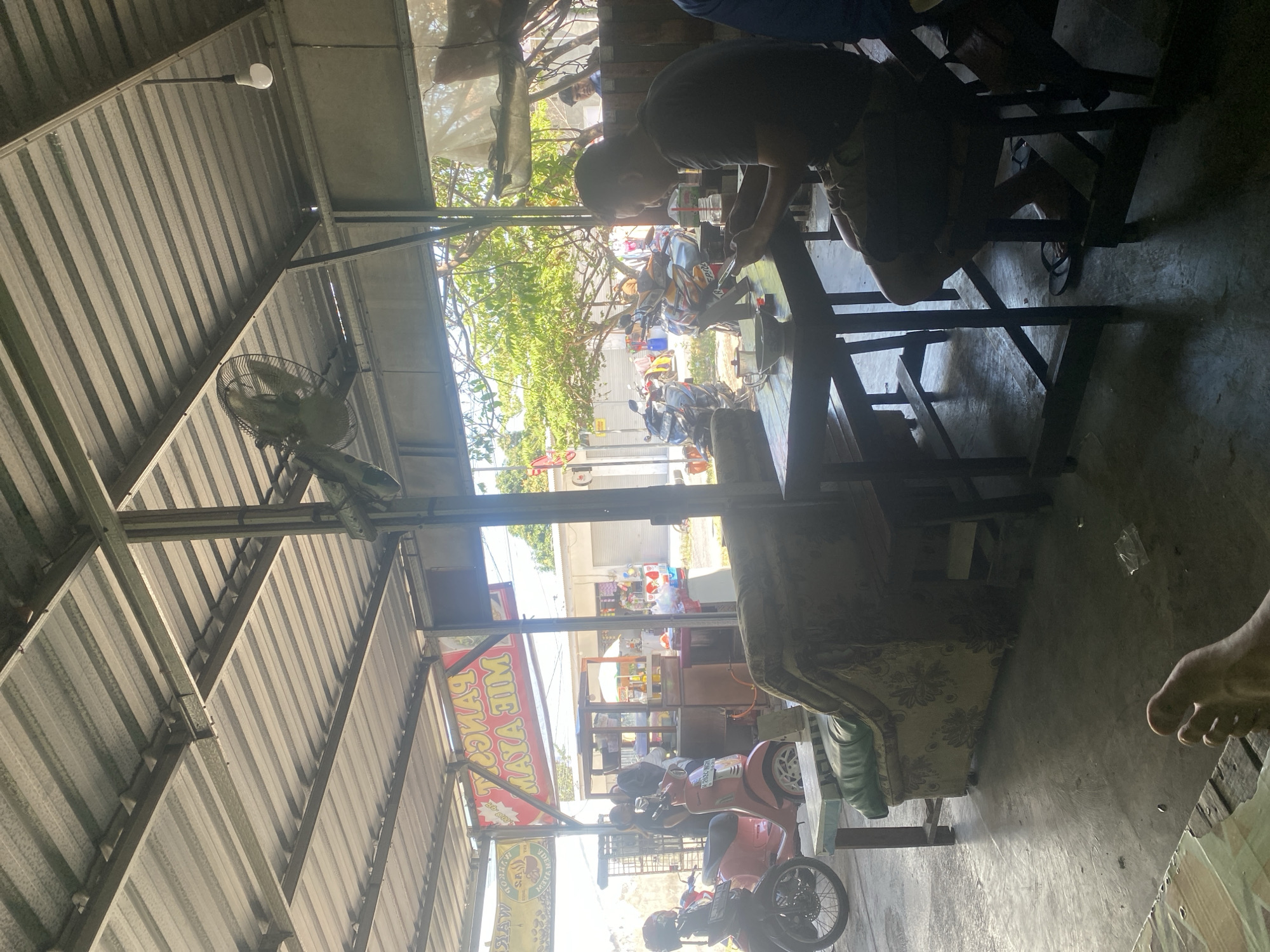 The image depicts an outdoor eating establishment in Karangbong, Indonesia, characterized by a corrugated metal roof supported by metal beams. A ceiling fan, a light bulb, and hanging signs are visible, one of which displays "PANGK MIE AYAM HIM". In the foreground, a man in a dark t-shirt and shorts is seated at a wooden table, engrossed in his smartphone. A bowl, a glass, and what appears to be food are present on the table. To the left, a pink scooter and a red motorcycle are parked. Further back, a floral-patterned sofa is positioned near another table. Several people are seen in the background near a food stall, and motorcycles are parked along the street. The ground is paved concrete, with some debris scattered around. Sunlight casts distinct shadows across the scene.