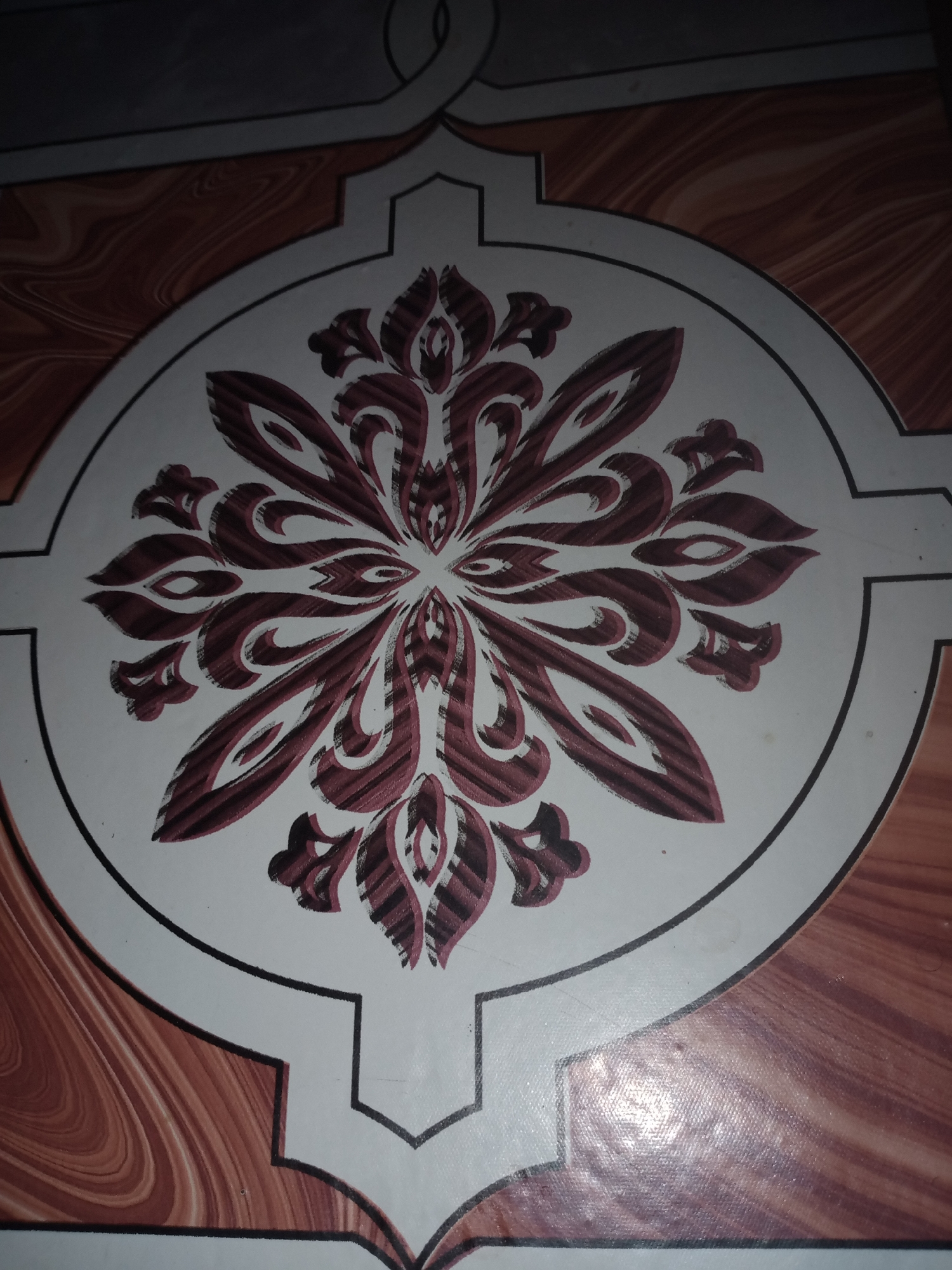 The image shows a close-up of a floor tile with a circular, symmetrical floral or paisley-like design in shades of dark red and white. The design has sharp, pointed elements radiating from a central point, with curved accents. The tile is surrounded by a border, and the floor itself appears to be made of tiles with a wood-grain pattern in reddish-brown and beige tones. The lighting suggests an indoor setting. There are no people or discernible actions visible. The image is taken from a high angle, looking down at the floor. No specific location in Jalingo, Nigeria can be identified from the image itself.